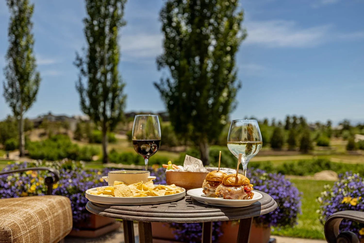 Outdoor table set with snacks, two glasses of wine (red and white), and mini sliders on a sunny day, with trees and a blue sky in the background.