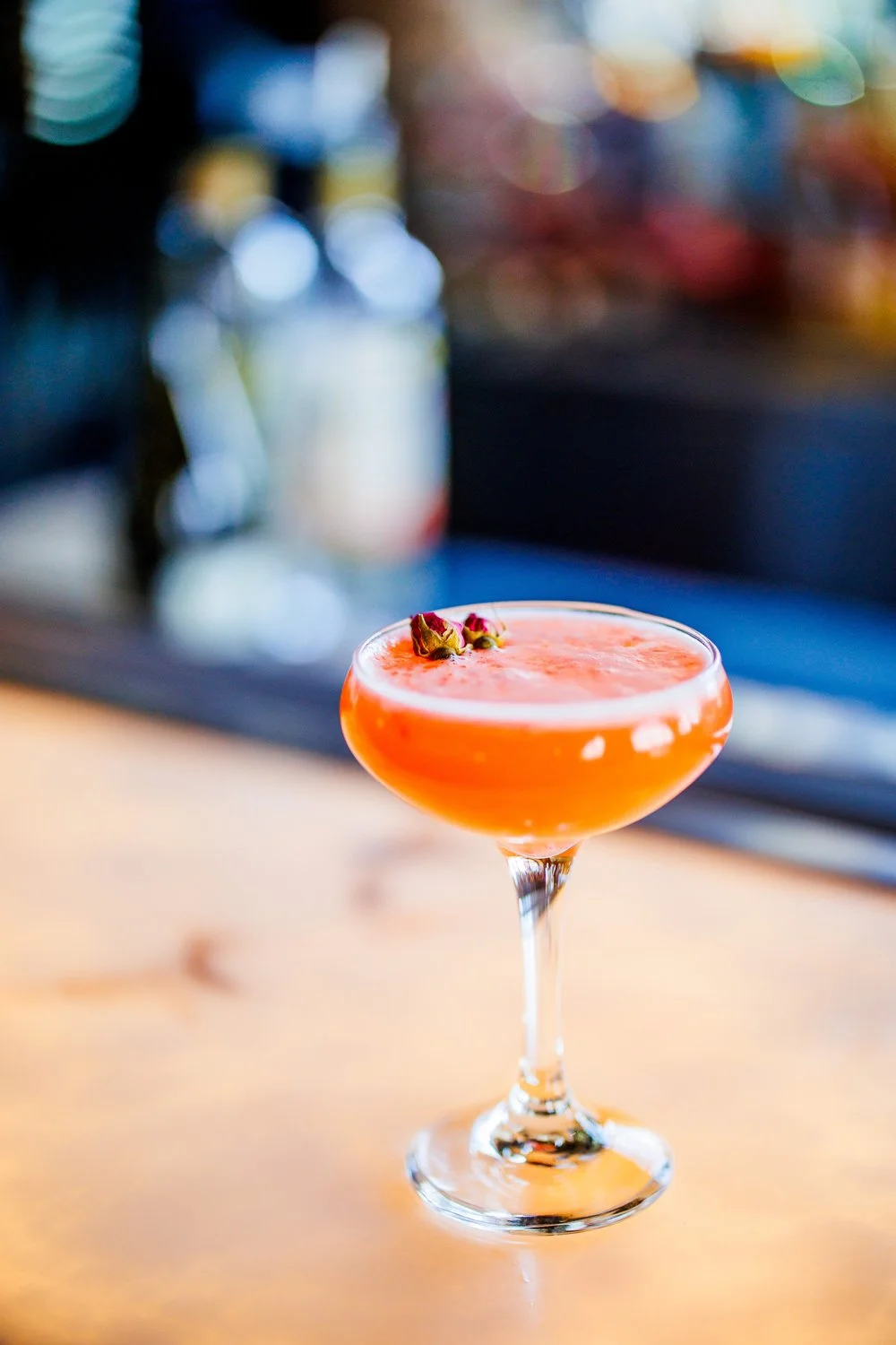 A cocktail in a coupe glass garnished with rosebuds, placed on a wooden bar counter with a blurred colorful background.