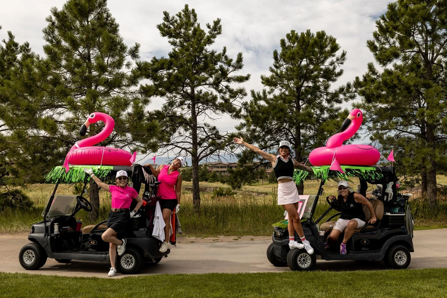 Four women celebrating on golf carts decorated with pink flamingo inflatables and green fringe, posing with arms raised and smiling outdoors with trees and sky in the background.