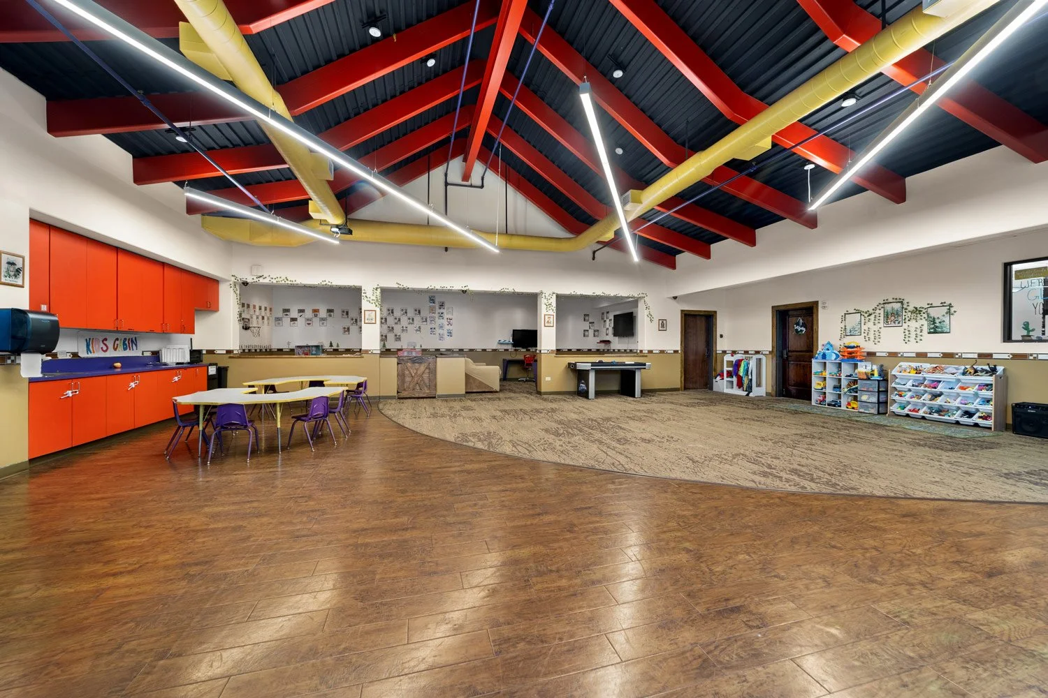 Empty activity room with wood flooring, red and yellow ceiling beams, and storage areas for children's toys and supplies in the Kids Cabin.
