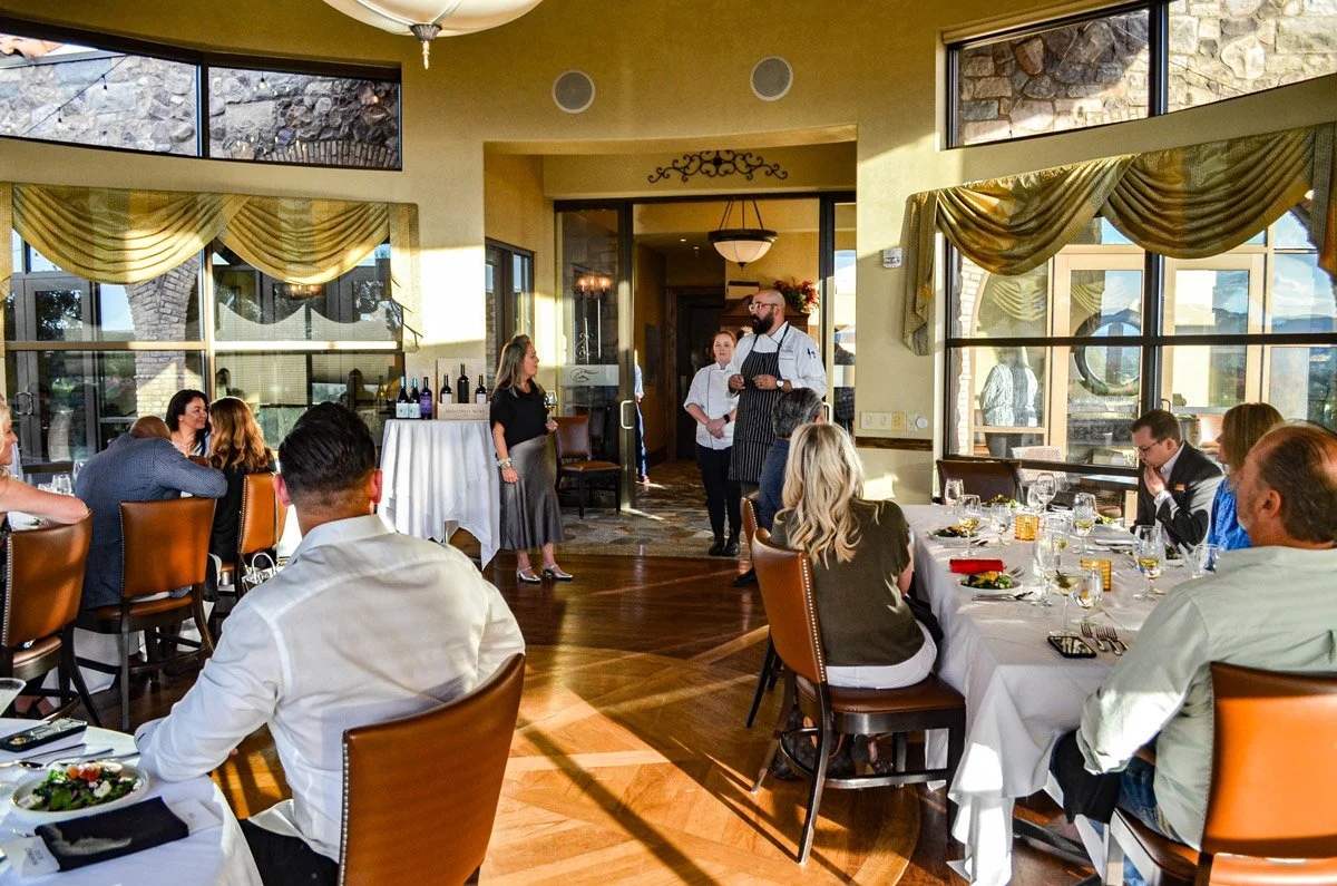 Guests seated at round tables in a restaurant or banquet hall, listening to a speaker and a chef standing by an open doorway, with wine bottles on a table behind them, during a daytime event with large windows and yellow walls.