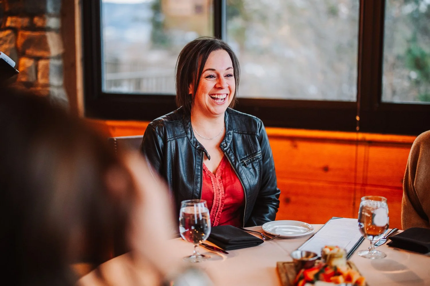A woman smiling and laughing seated at a restaurant table with a glass of water, plates, and food, near a large window with a view of trees outside.