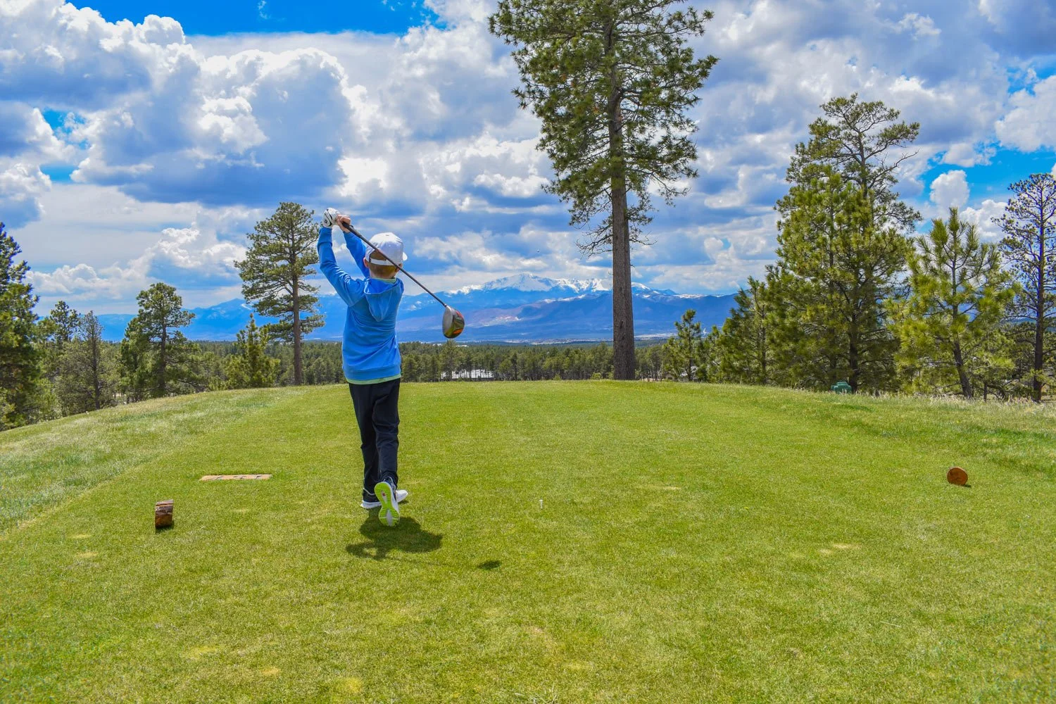 Child in blue jacket and white cap playing golf on a green golf course with trees and mountains in the background under a partly cloudy sky.