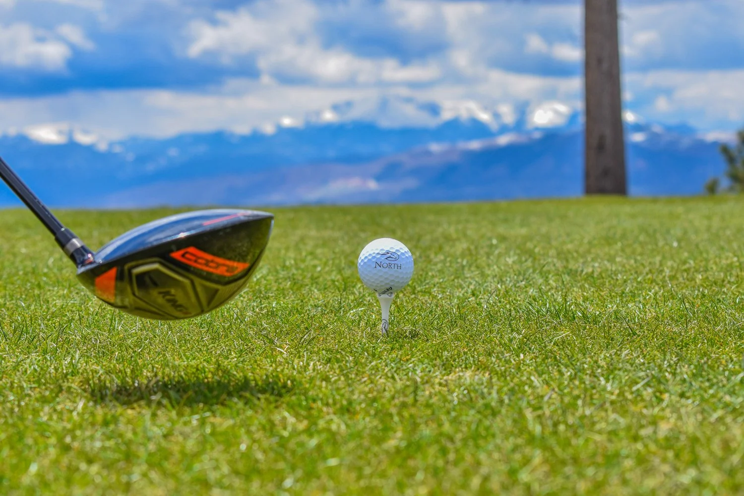 Golf club near a golf ball on a tee on the grass with mountains and blue sky in the background.