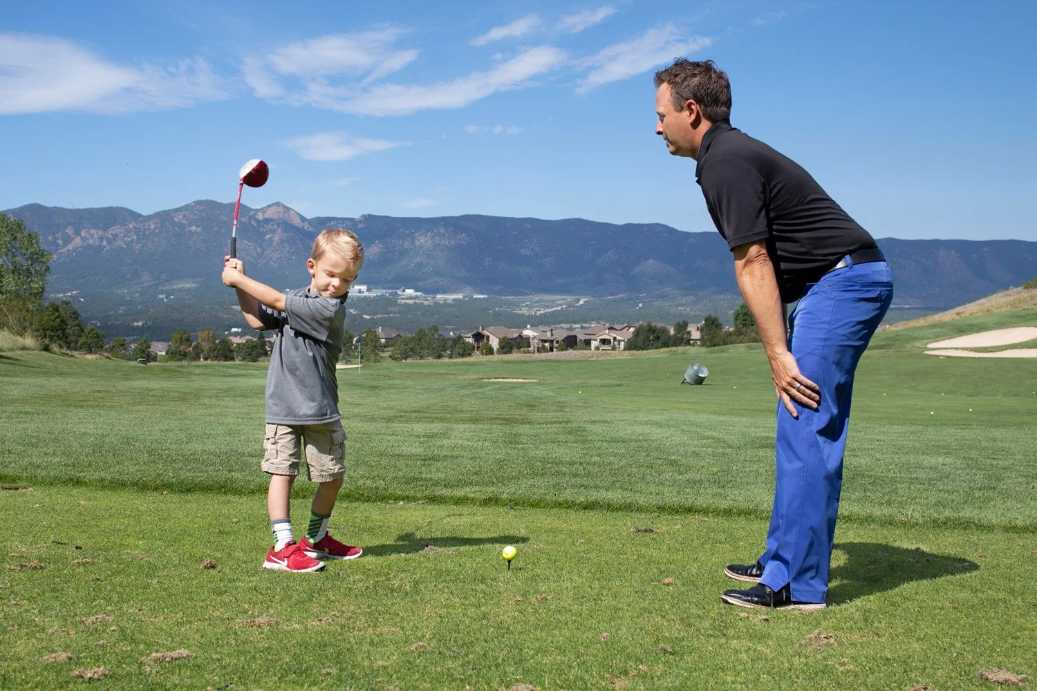 A young boy practicing golf with his instructor, on a golf course with mountains in the background. The boy is holding a golf club, preparing to hit a golf ball, while the man is bent over, watching him.