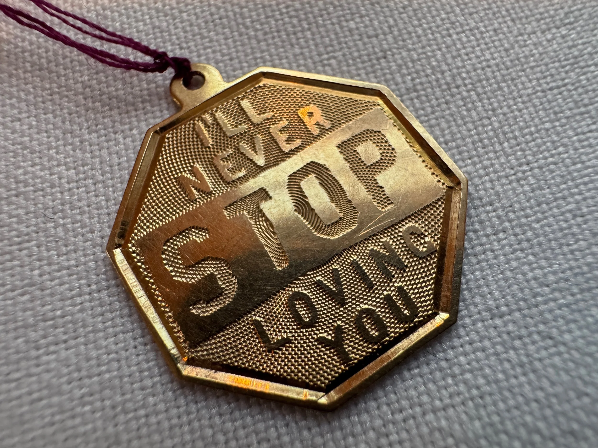 Gold-colored badge with the words 'NSU' and 'Land' engraved on its textured surface, with a purple string attached at the top, lying on gray fabric.