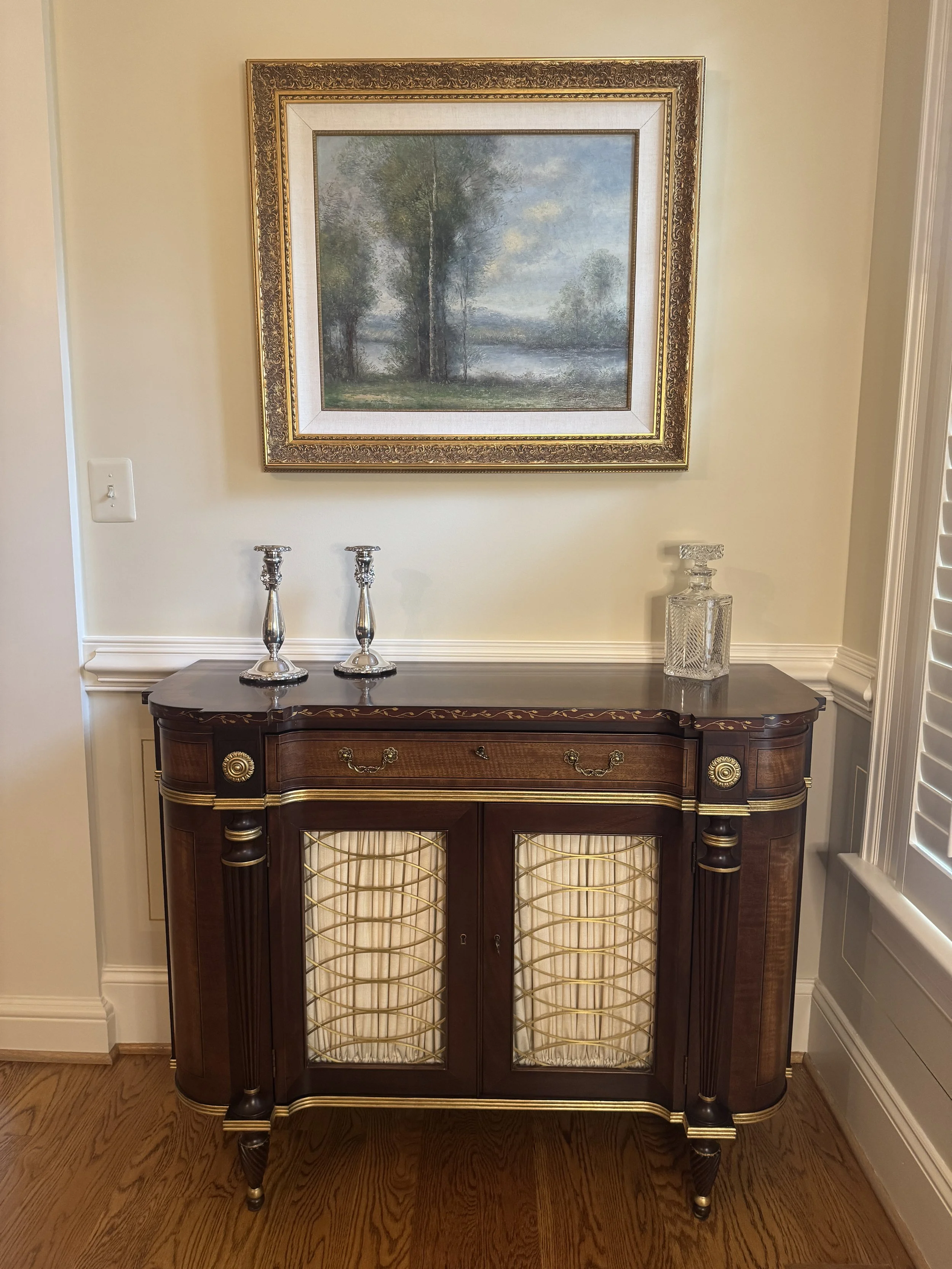 A vintage wooden sideboard with gold accents and lattice glass doors, topped with two silver candlesticks and a crystal decanter, hanging on a beige wall below a landscape painting of trees and a river.