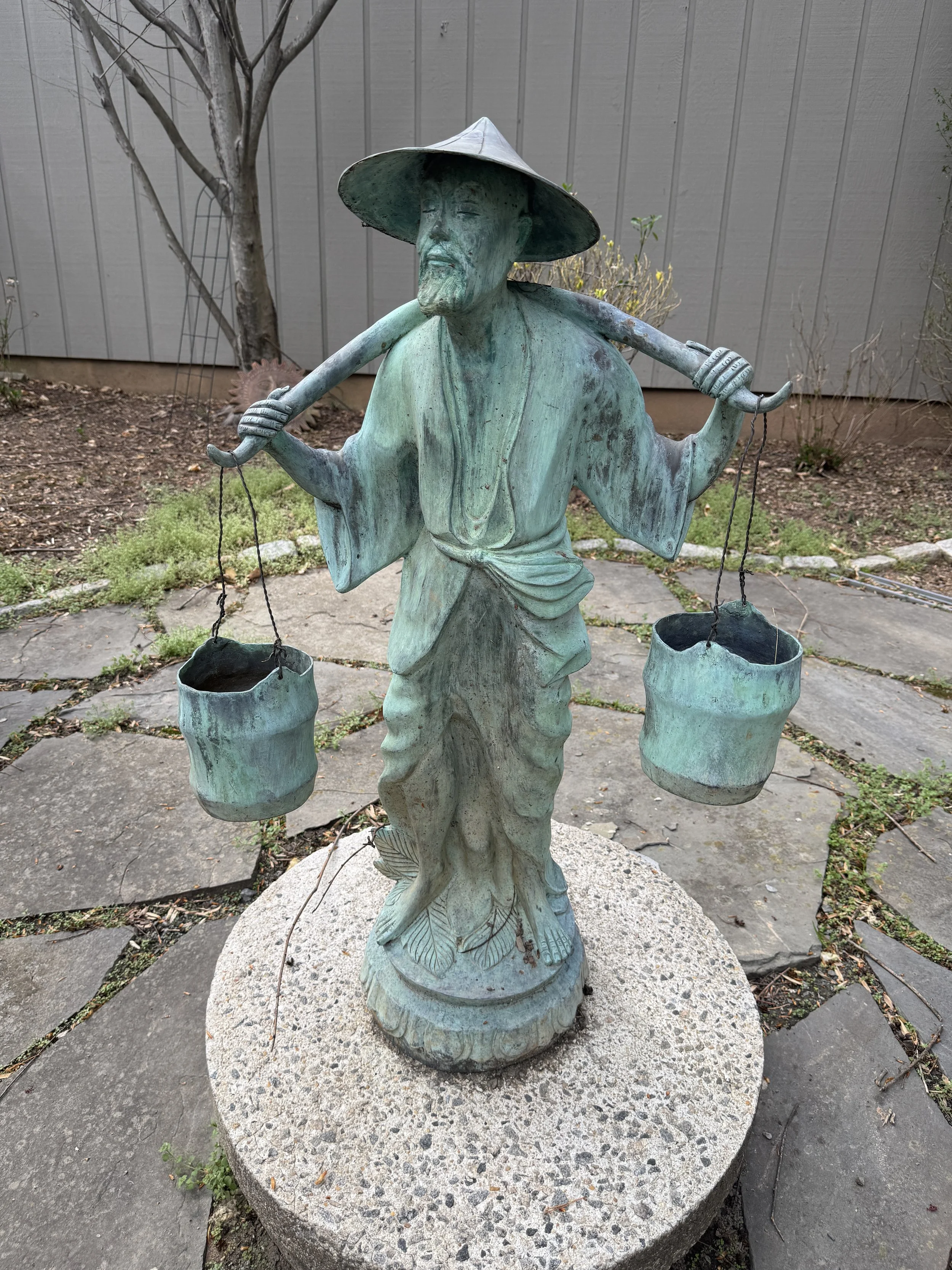 Bronze statue of a man in traditional clothing with a wide hat, carrying two buckets on a pole across his shoulders, standing on a concrete base in a garden.