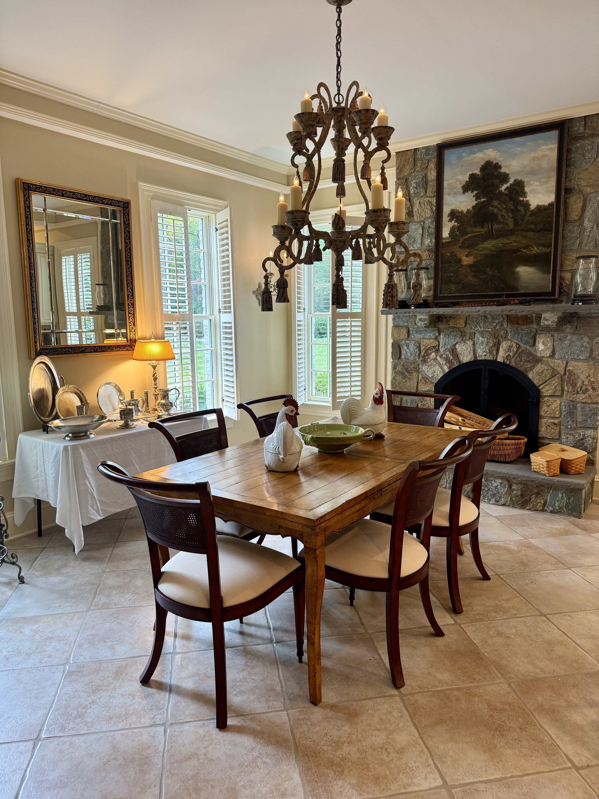 A cozy dining room with a wooden table, six chairs, a stone fireplace, a chandelier, and a side table with decorative items. Windows with shutters let in natural light.