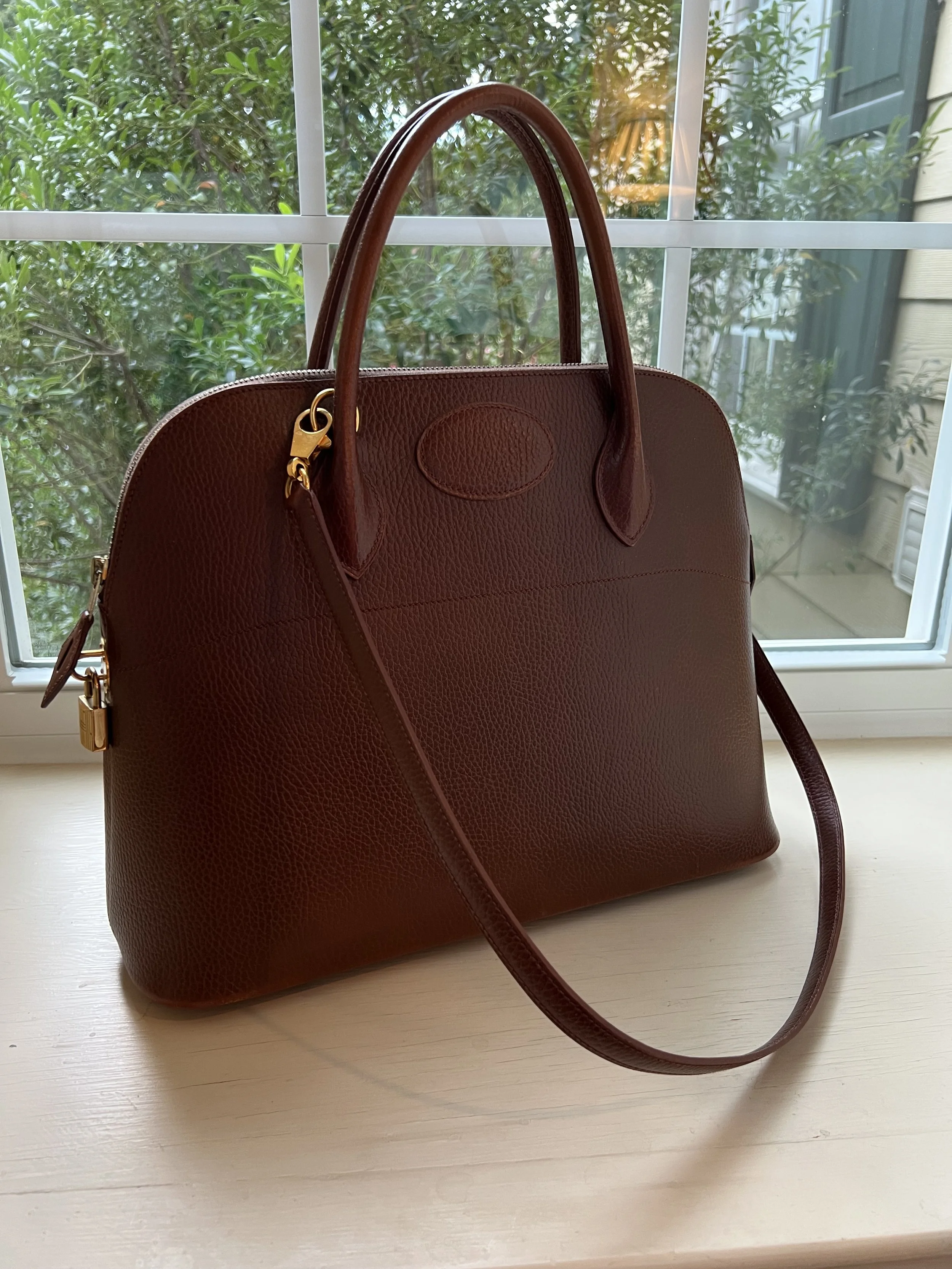 Brown leather handbag on a white surface in front of a window with greenery outside.