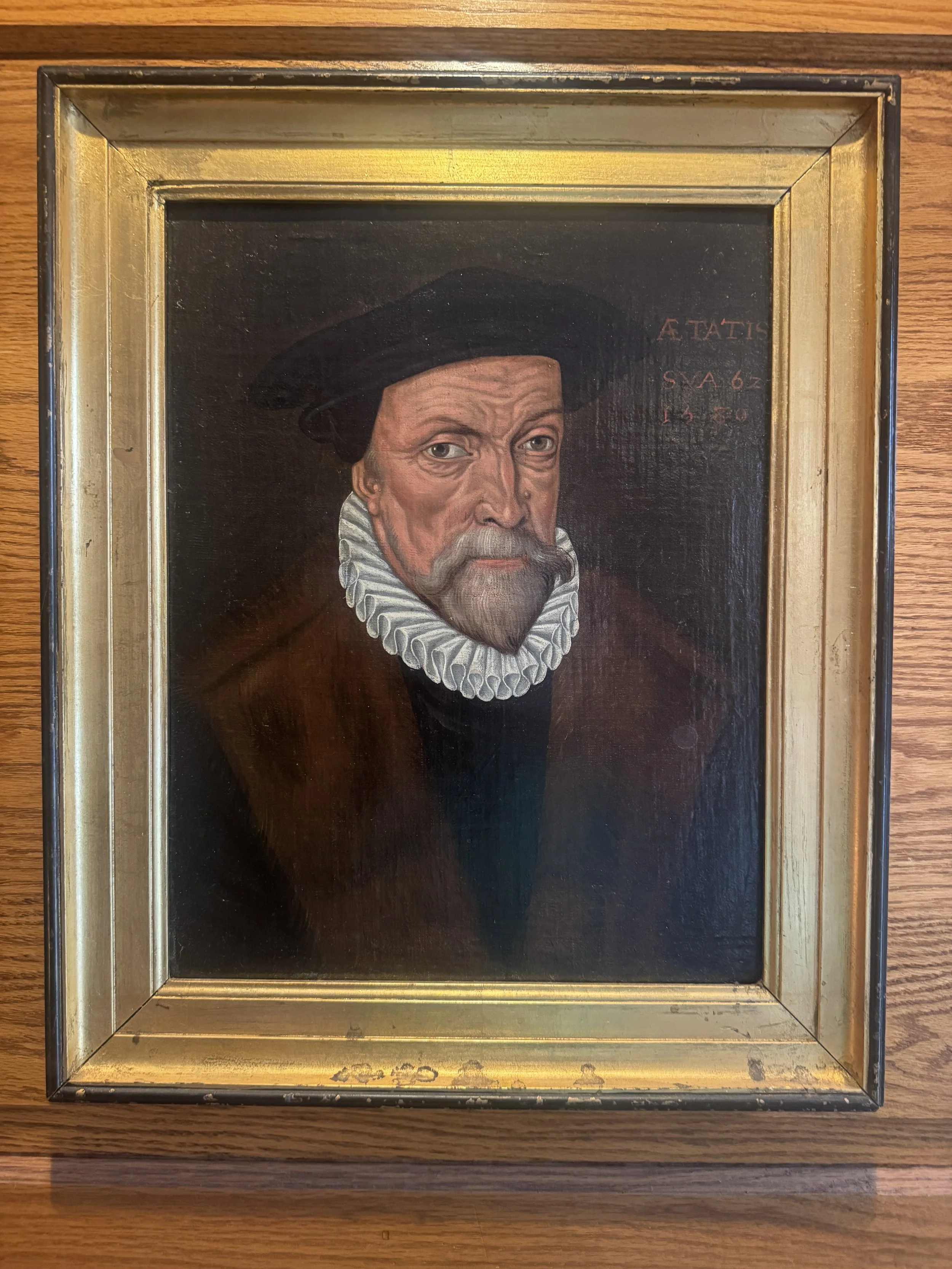 Painting of a man in historical attire with a ruffled collar and a black hat, with text in Latin that appears to say 'A TATIS' and other words partially visible.