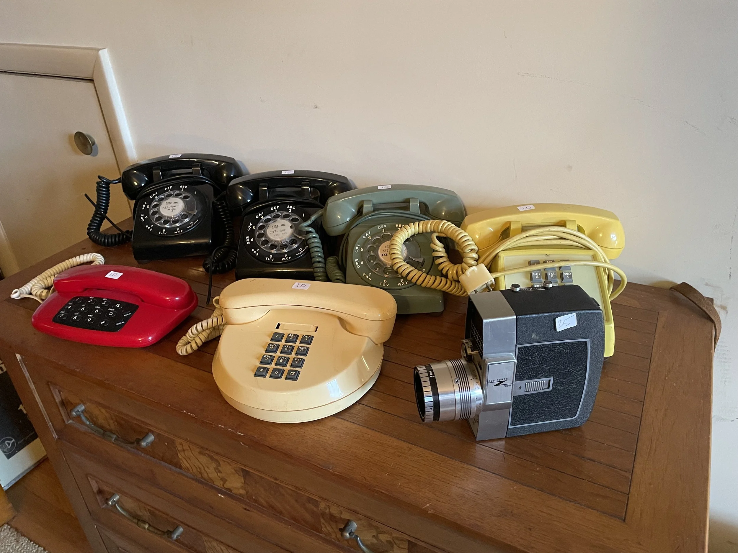 Collection of vintage rotary and push-button telephones of various colors, including black, green, yellow, beige, and red, and a vintage box camera on a wooden table.