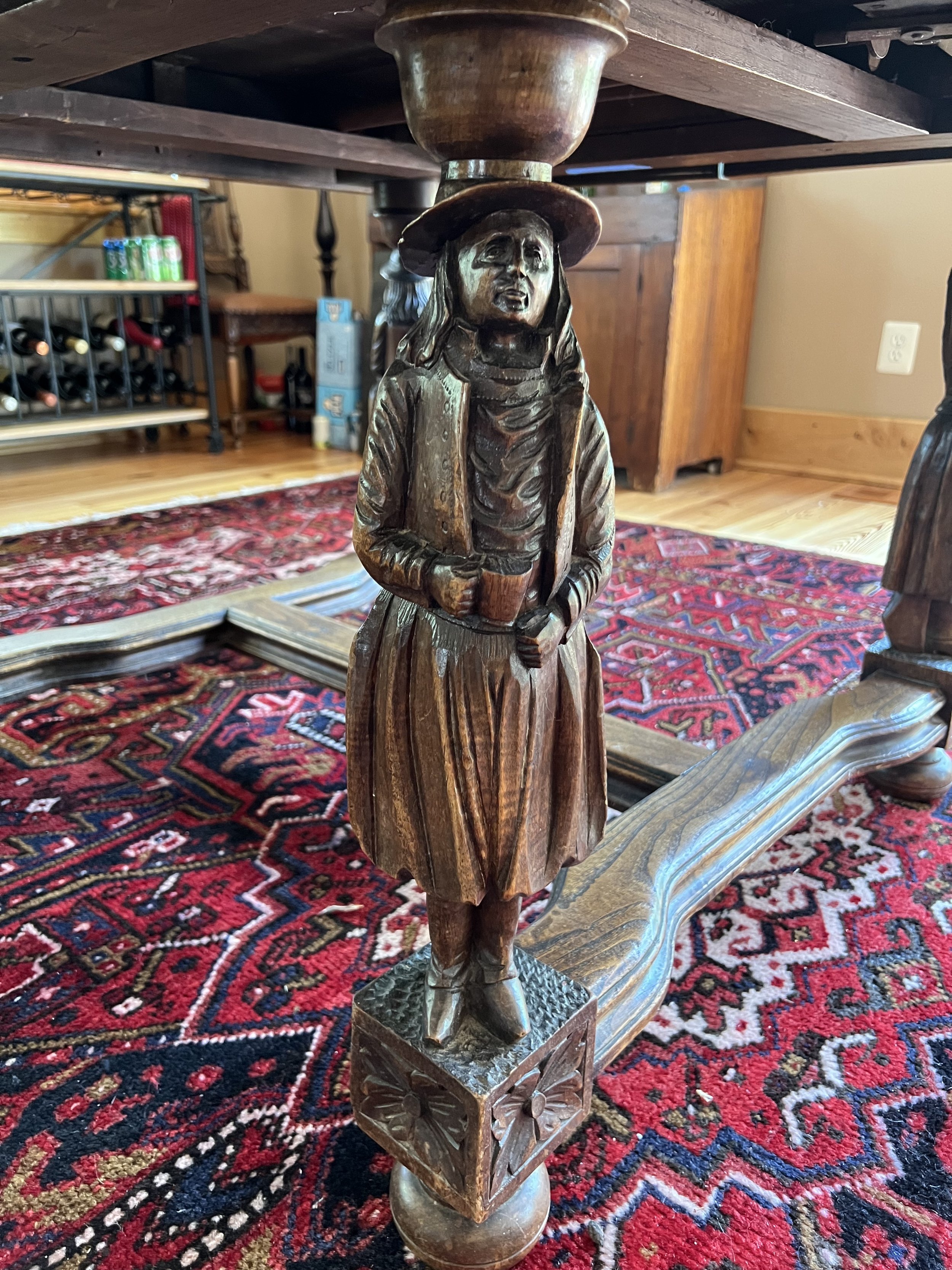 Wooden carved statue of a woman holding a book and wearing a hat, as part of a table leg, set on a decorative red and black patterned rug.