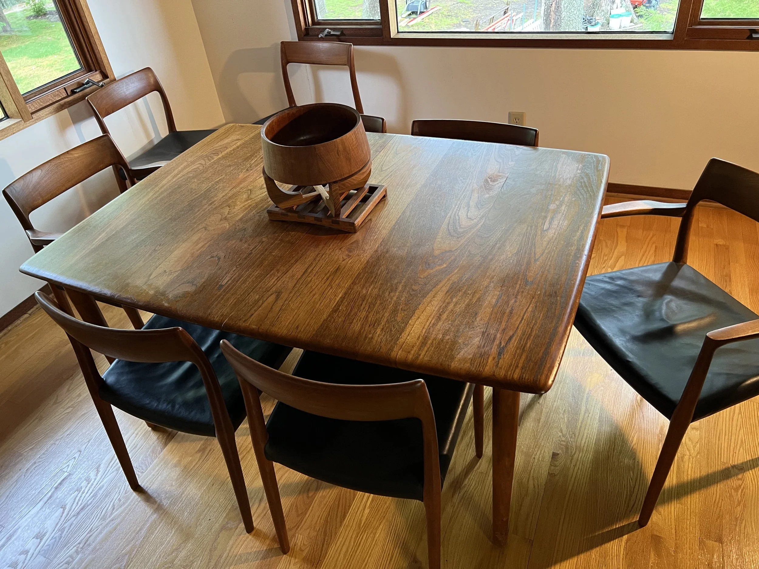 Wooden dining table with six black-cushioned chairs around it, set in a room with large windows and a wooden floor, with a decorative bowl on a wooden stand on the table.