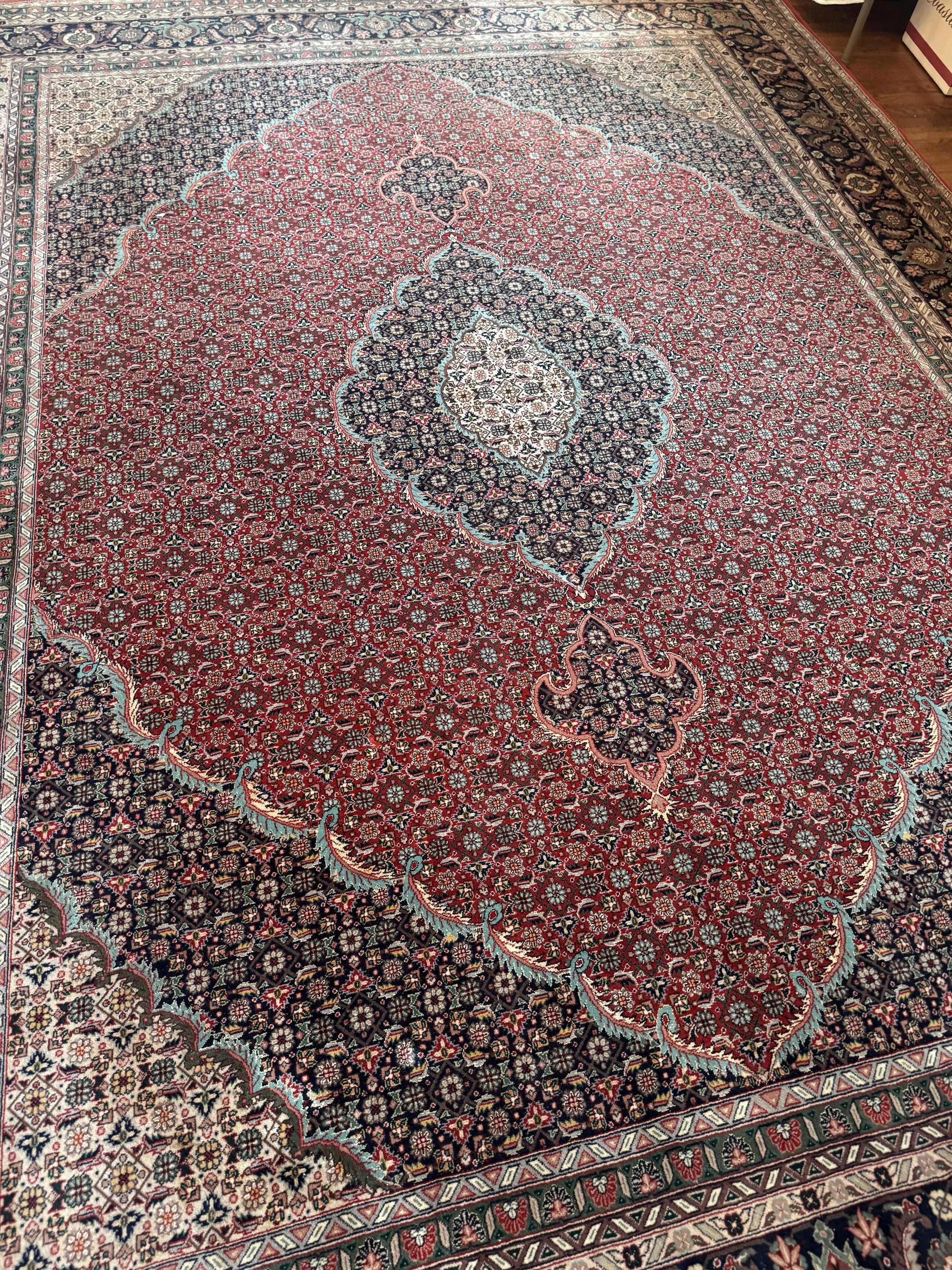 A large, traditional Persian-style rug with intricate floral patterns in red, black, cream, and blue colors, featuring a central medallion design and decorative border.