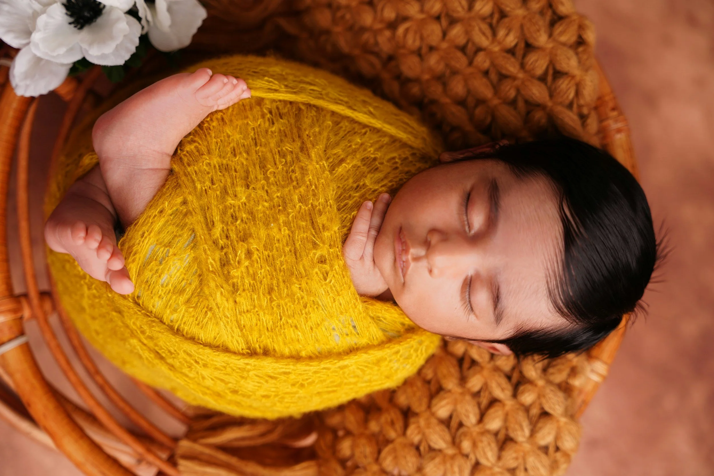 A sleeping baby wrapped in a yellow knitted blanket, lying on a woven basket with white flowers nearby.
