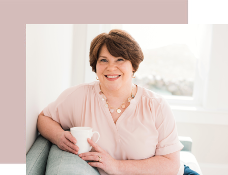 A woman with short brown hair smiling and holding a white coffee mug, sitting on a sofa near a window.