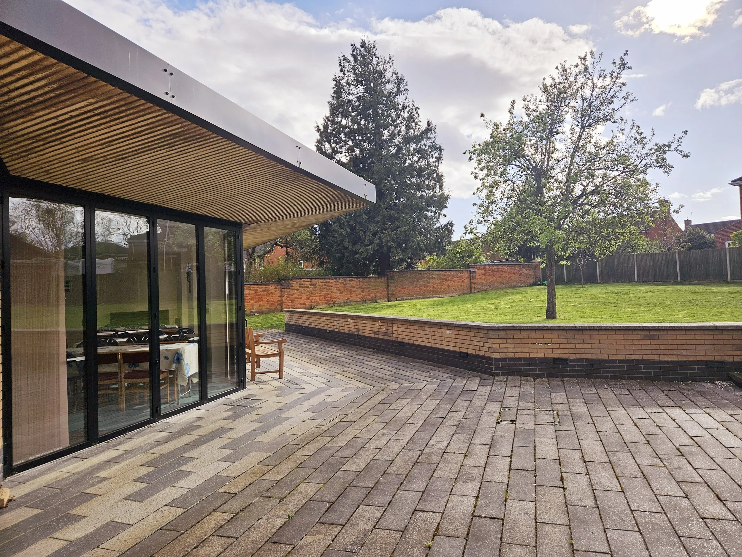 A modern house with glass sliding doors, a wooden ceiling overhang, a patio with brick paving, and a grassy yard with trees enclosed by a brick wall and wooden fence.