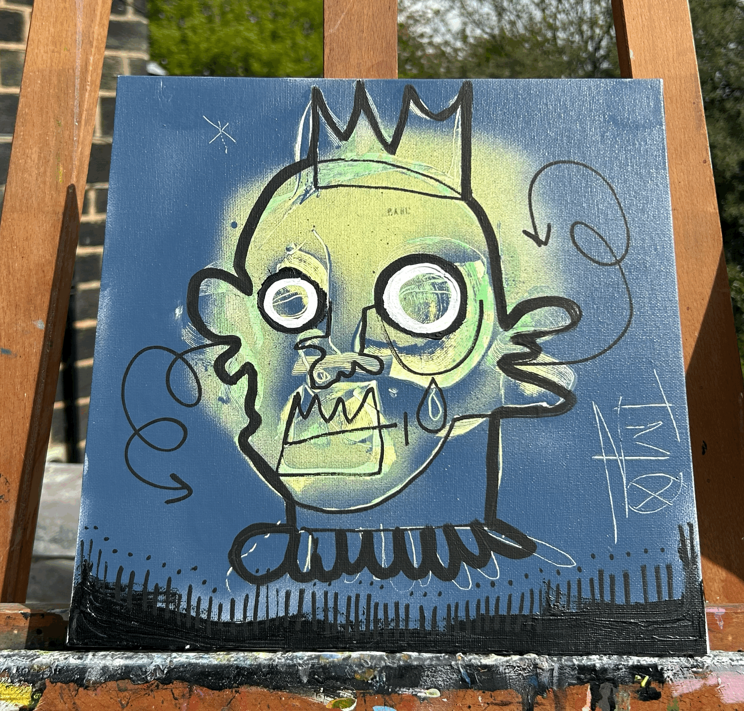 Street art painting of a cartoonish, ghostly figure with a crown, large circular eyes, sharp teeth, and a tear, with scribbles and signatures around it, on a blue background.