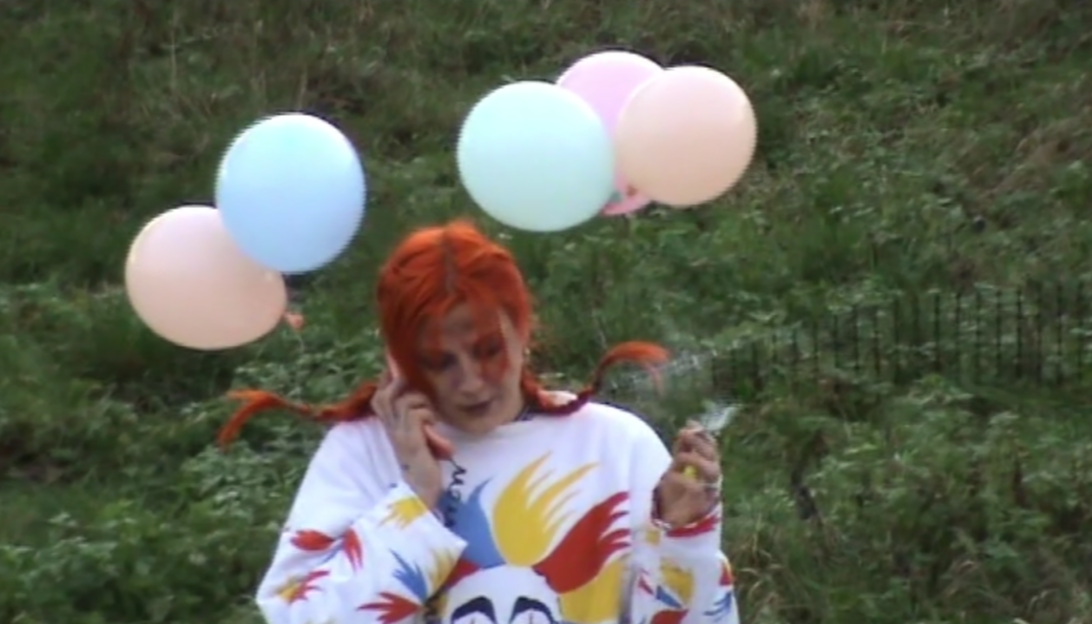 A woman talking on a cellphone outdoors with colorful balloons behind her and a grassy background.