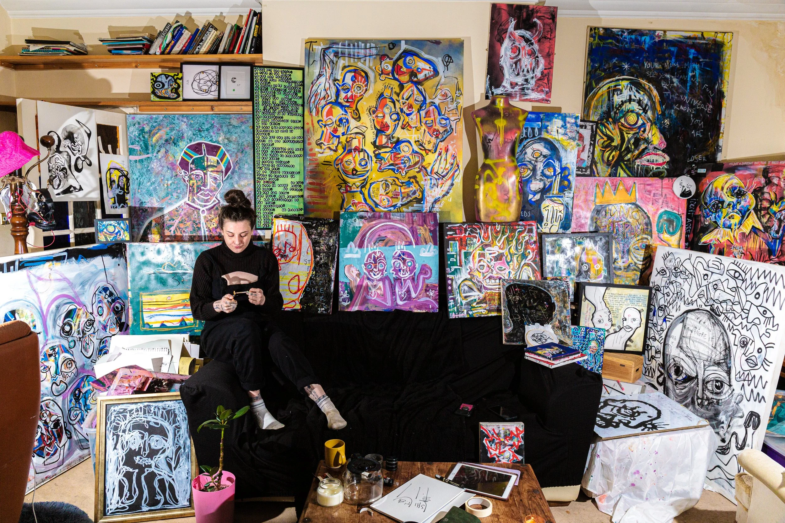 Artist working in a cluttered studio surrounded by colorful abstract paintings and sketches, with art supplies and a tablet on the table.