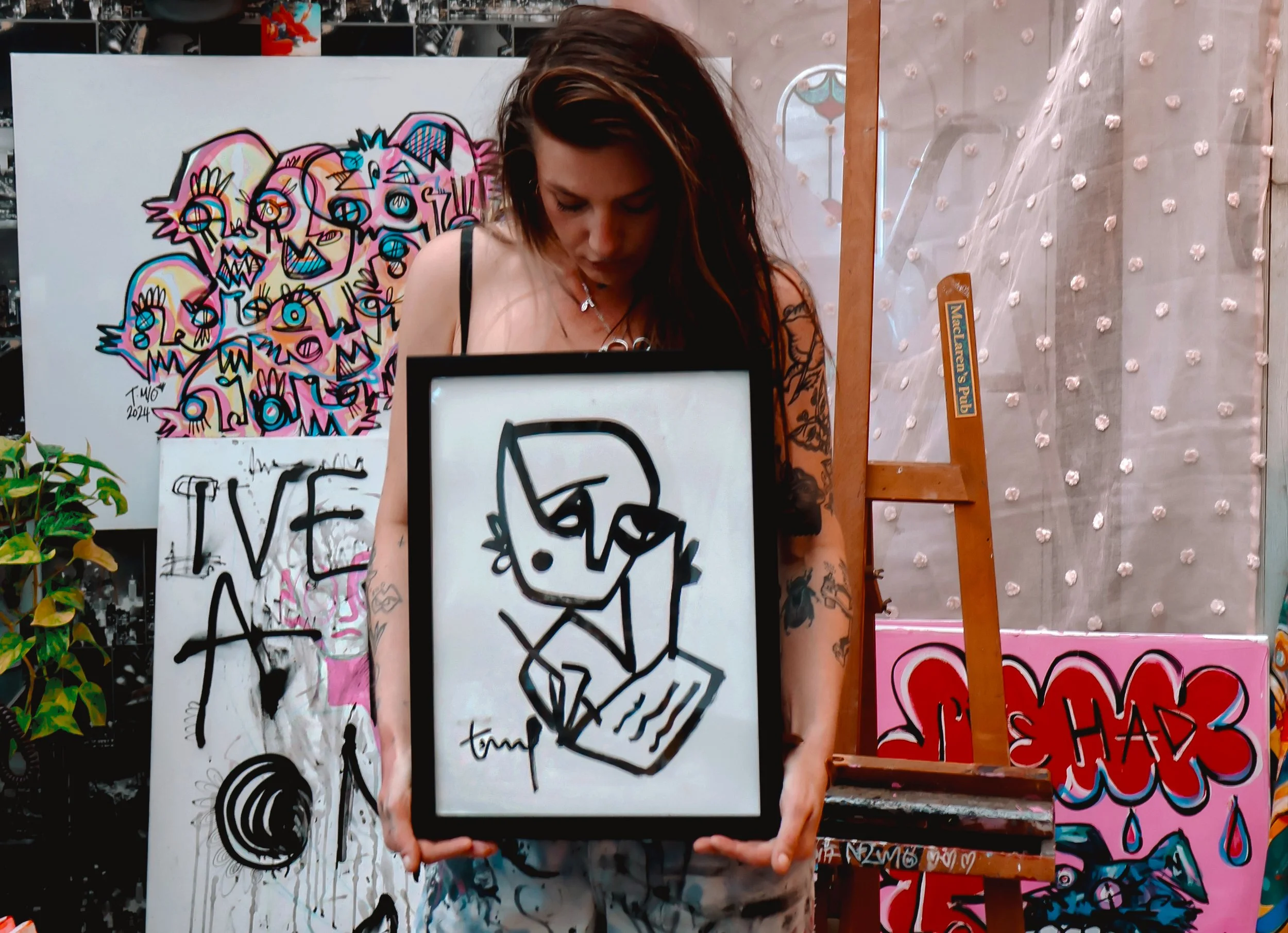 Young woman with tattoos and dark hair holding a framed abstract black and white drawing. Behind her are colorful graffiti-style paintings and street art in an artist's studio or outdoor setting.