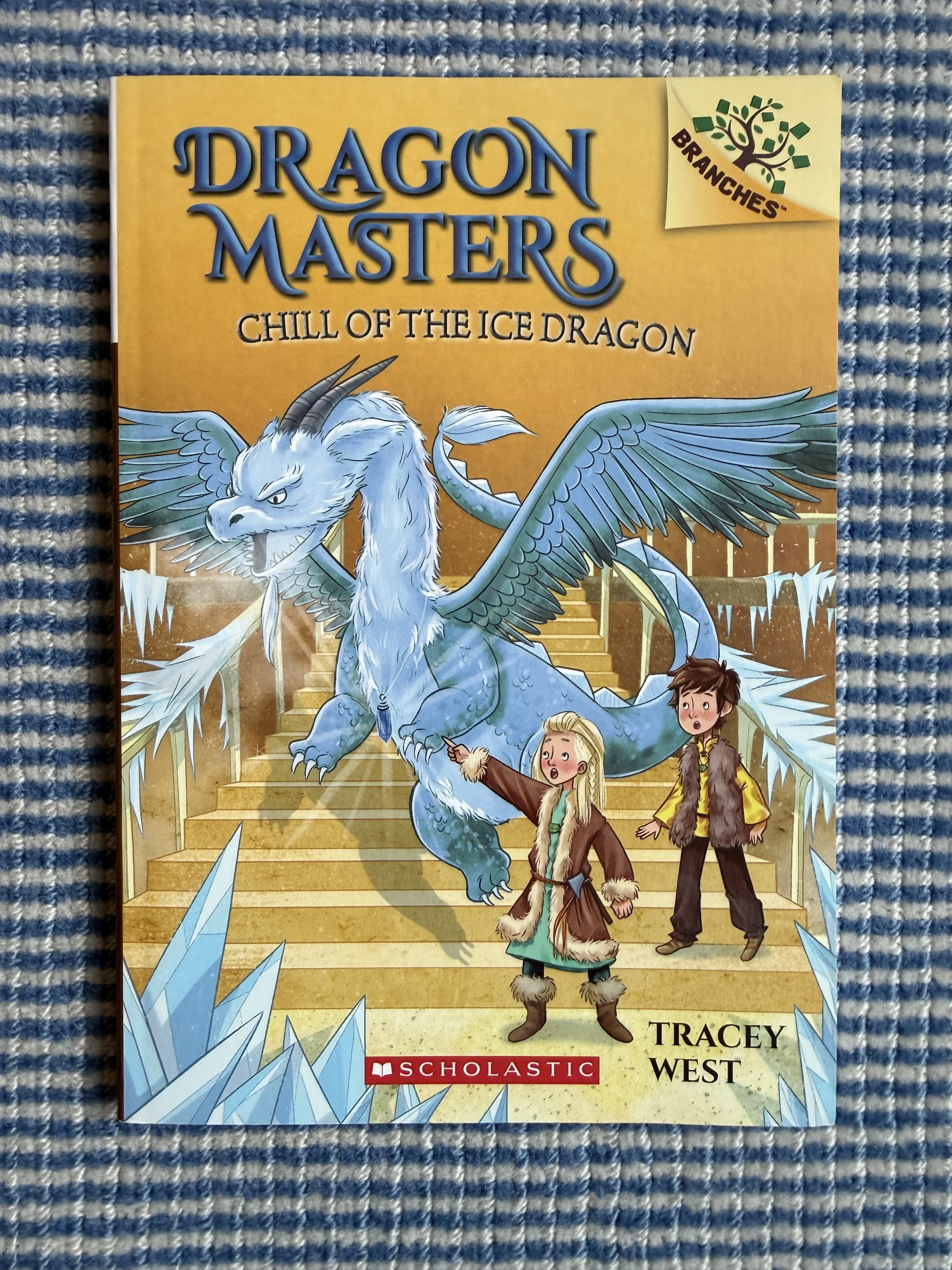 Book cover of 'Dragon Masters: Chill of the Ice Dragon' by Tracey West, featuring an illustration of a large blue ice dragon with two children in fur-lined clothing standing on a staircase, surrounded by ice shards. Publisher logo of Scholastic is at the bottom.