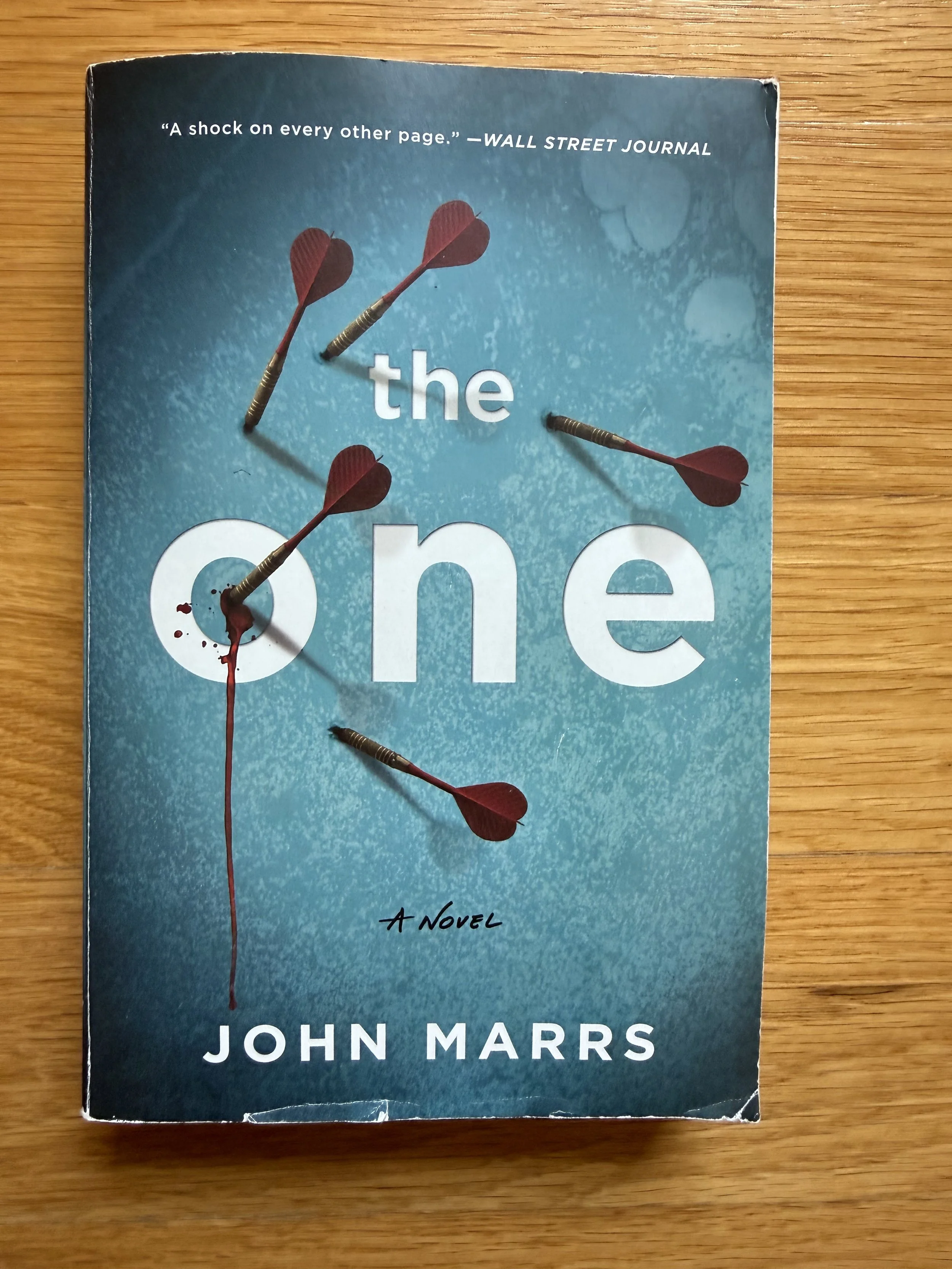 A book titled 'The One' by John Marrs with three darts stuck in the cover, one of which is bleeding red ink or paint.