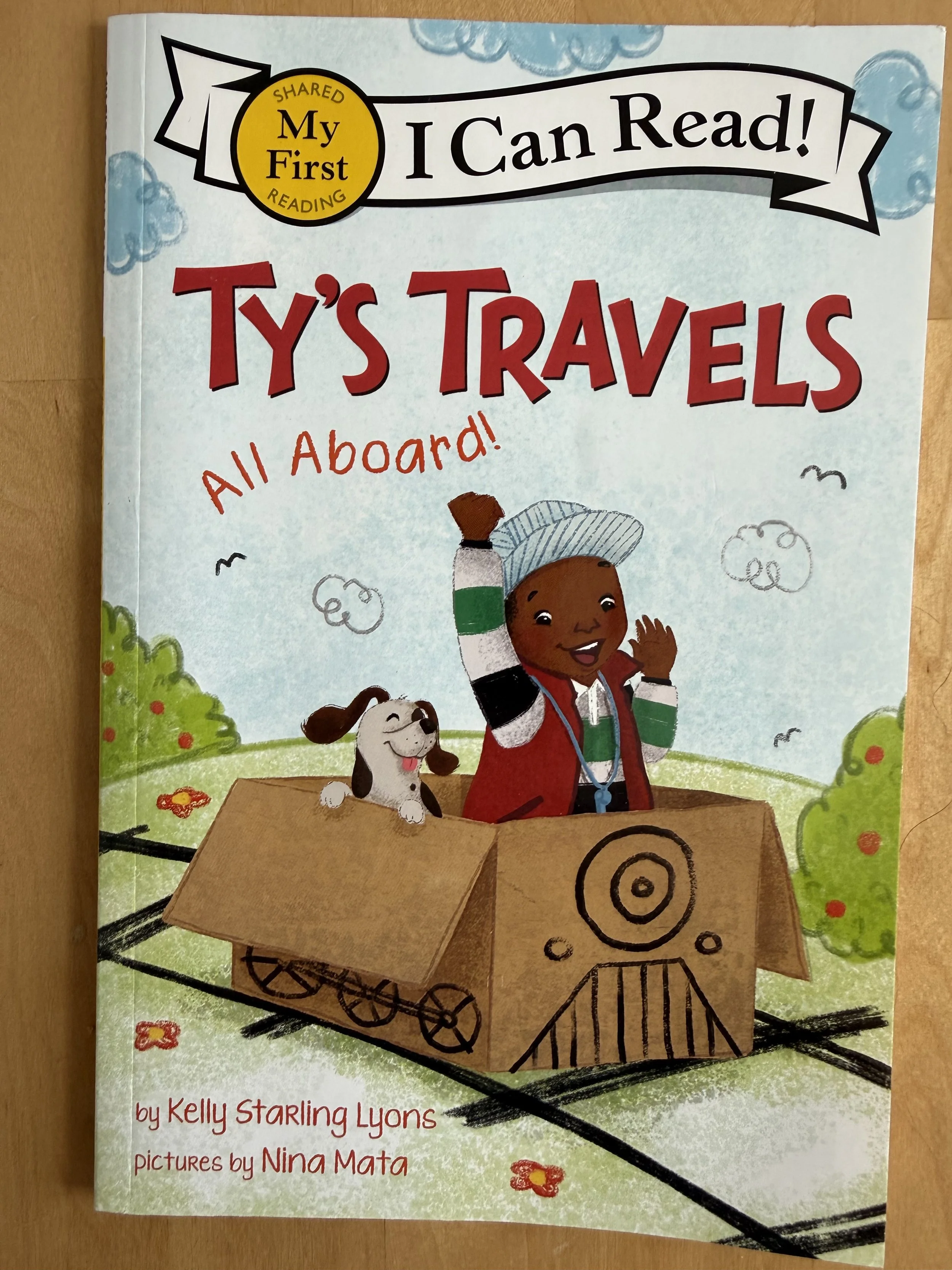 Cover of a children's book titled "My First I Can Read! Ty's Travels." The cover features an illustration of a smiling child with dark skin and short curly hair, wearing a colorful jacket and a hat, waving from inside a cardboard box on train tracks. A happy dog with floppy ears peers out of the box. There are fluffy clouds and green bushes, with red flowers scattered around.