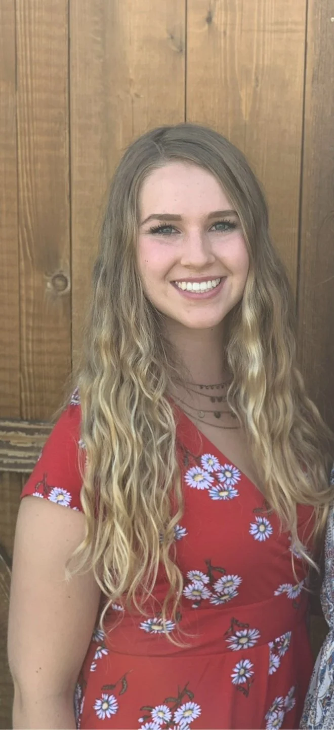 binge eating coach Amber Abila with long, wavy blonde hair wearing a red dress with white daisy flower print, smiling at the camera, standing in front of a wooden wall.