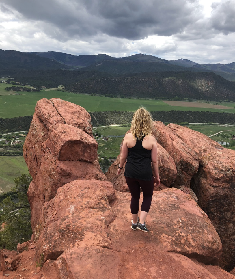 binge eating coach Amber Abila in black athletic clothing with curly blonde hair walking on red rock formations overlooking a green valley with mountains in the background under a cloudy sky.