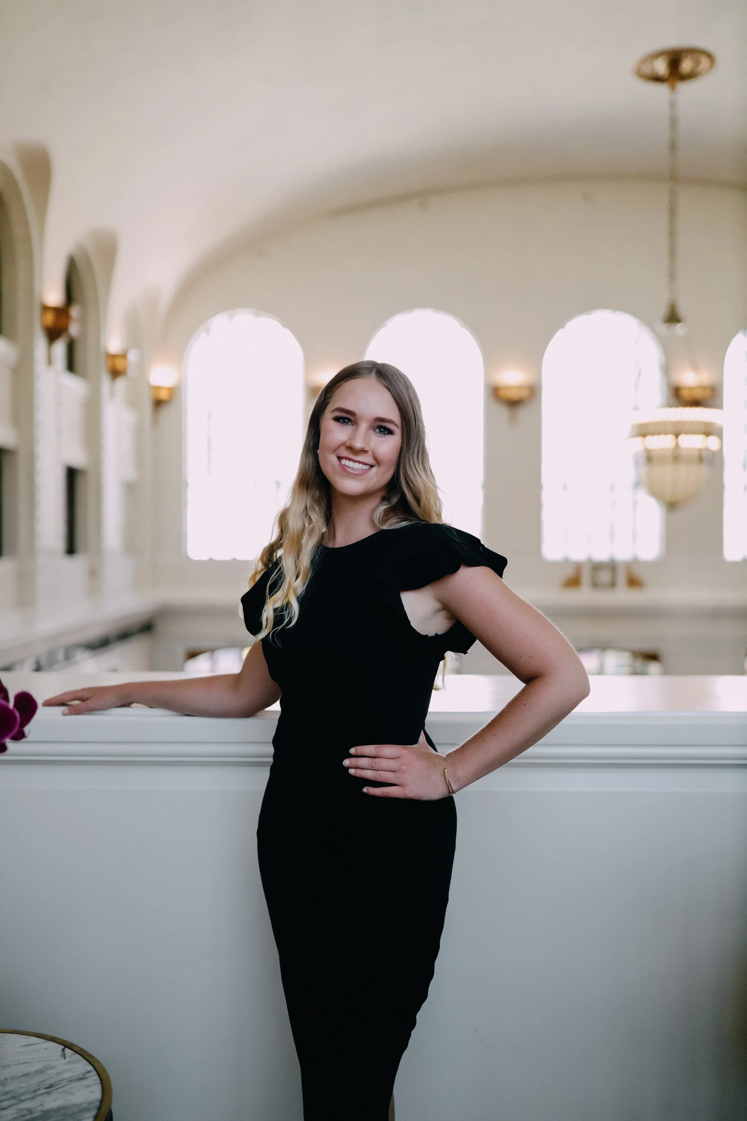 A young woman with long wavy blonde hair smiling, wearing a black dress, standing in a bright room with large arched windows and elegant lighting fixtures.