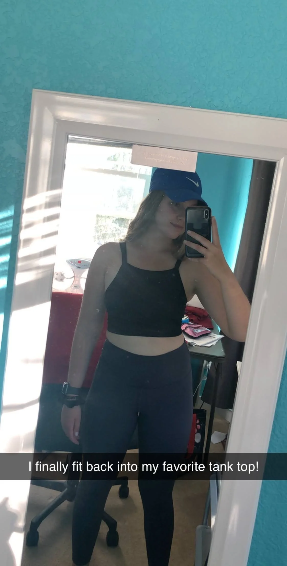 binge eating coach Amber Abila taking a mirror selfie showing her athletic outfit, wearing a black tank top and black leggings, with a blue Nike cap. She is in a room with a turquoise wall and a window with sunlight.
