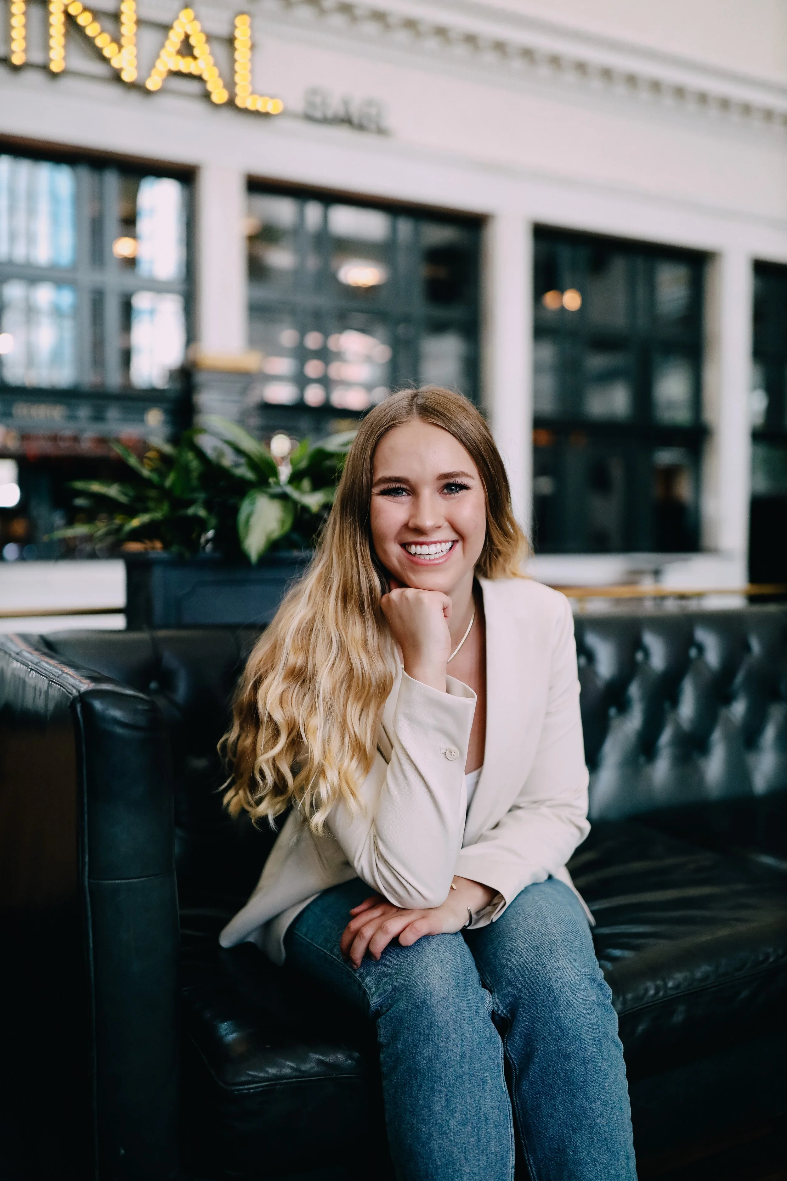 A woman with long, wavy blonde hair sitting on a black leather couch in a modern indoor setting, smiling at the camera with her chin resting on her hand, wearing a white blazer and blue jeans, with a large green plant and cityscape visible through windows in the background.