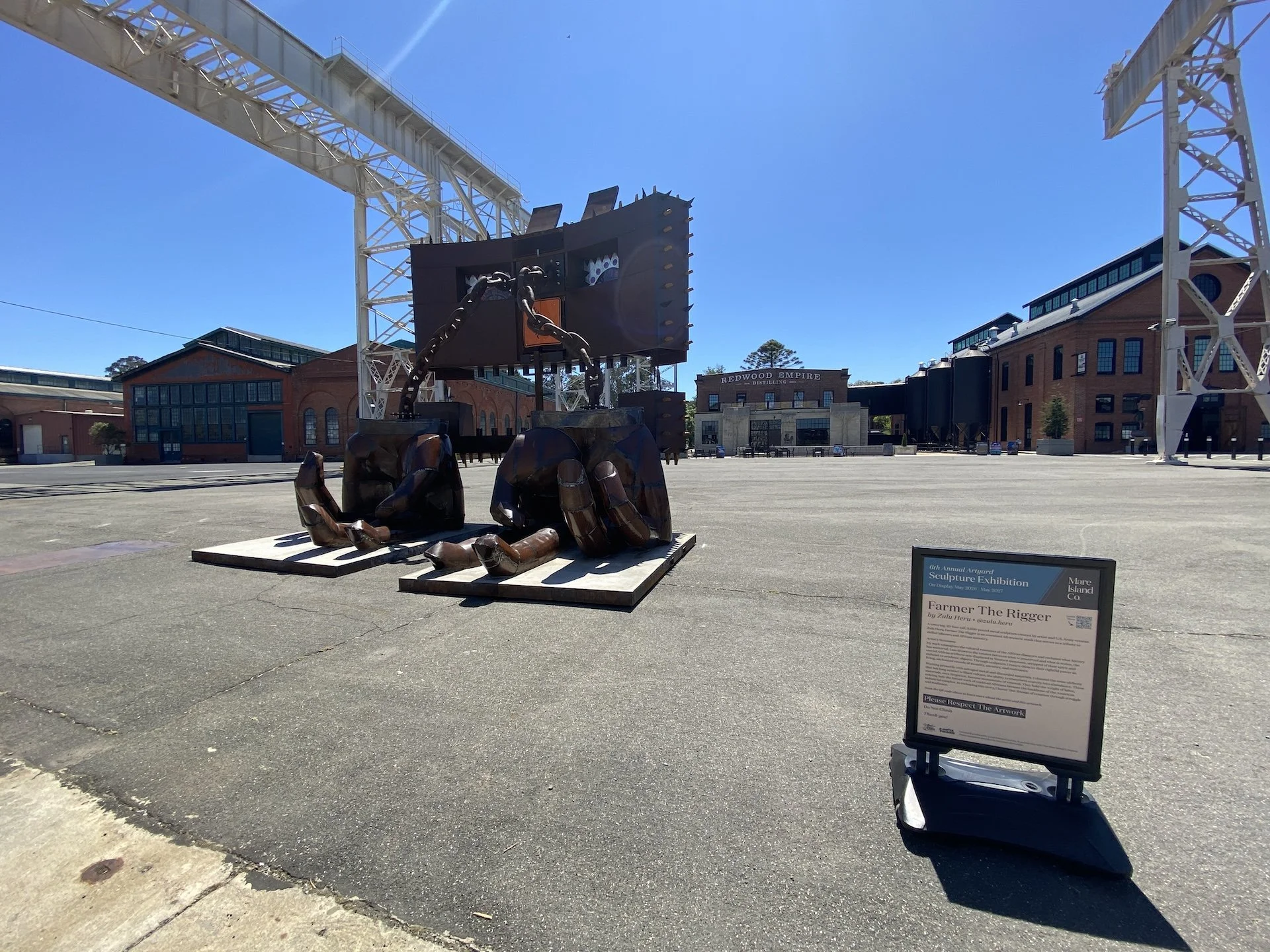Sculpture titled 'Farmer The Rigger' displayed outdoors on a platform, featuring large, rust-colored mechanical hands and chains. In the background, there are brick buildings, a large digital screen, and a sign for the Redwood Empire Distilling Compa