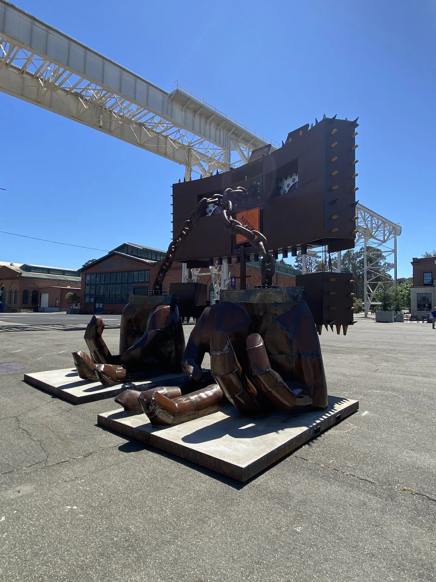 Outdoor sculpture of a large mechanical tiger with a chain-link chain around its neck, situated on a platform in an open parking lot with buildings in the background and a blue sky overhead.