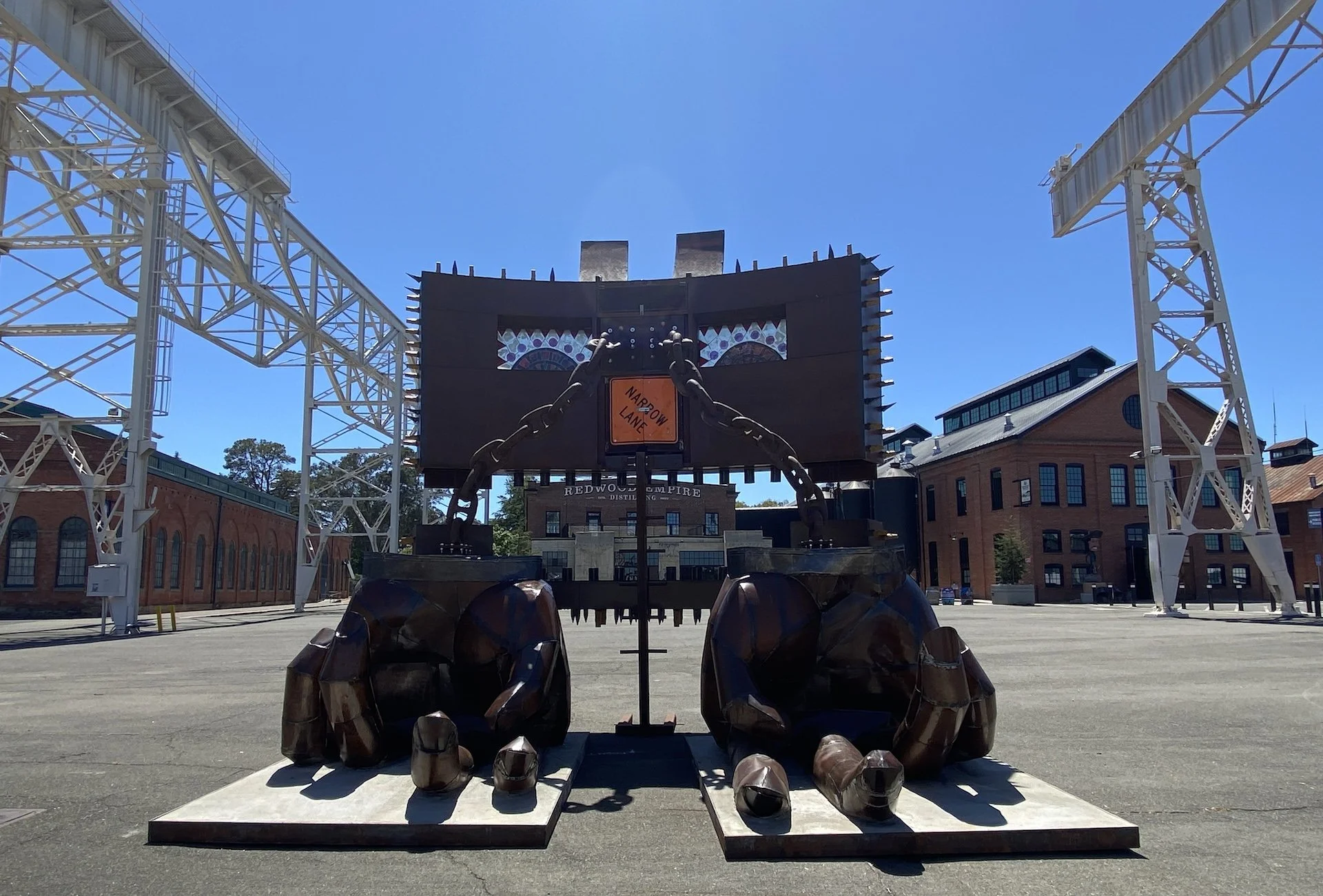 Large outdoor sculpture of a bear made of rusted metal, with a chain attached, standing against a backdrop of a blue sky, metal structures, and brick buildings.