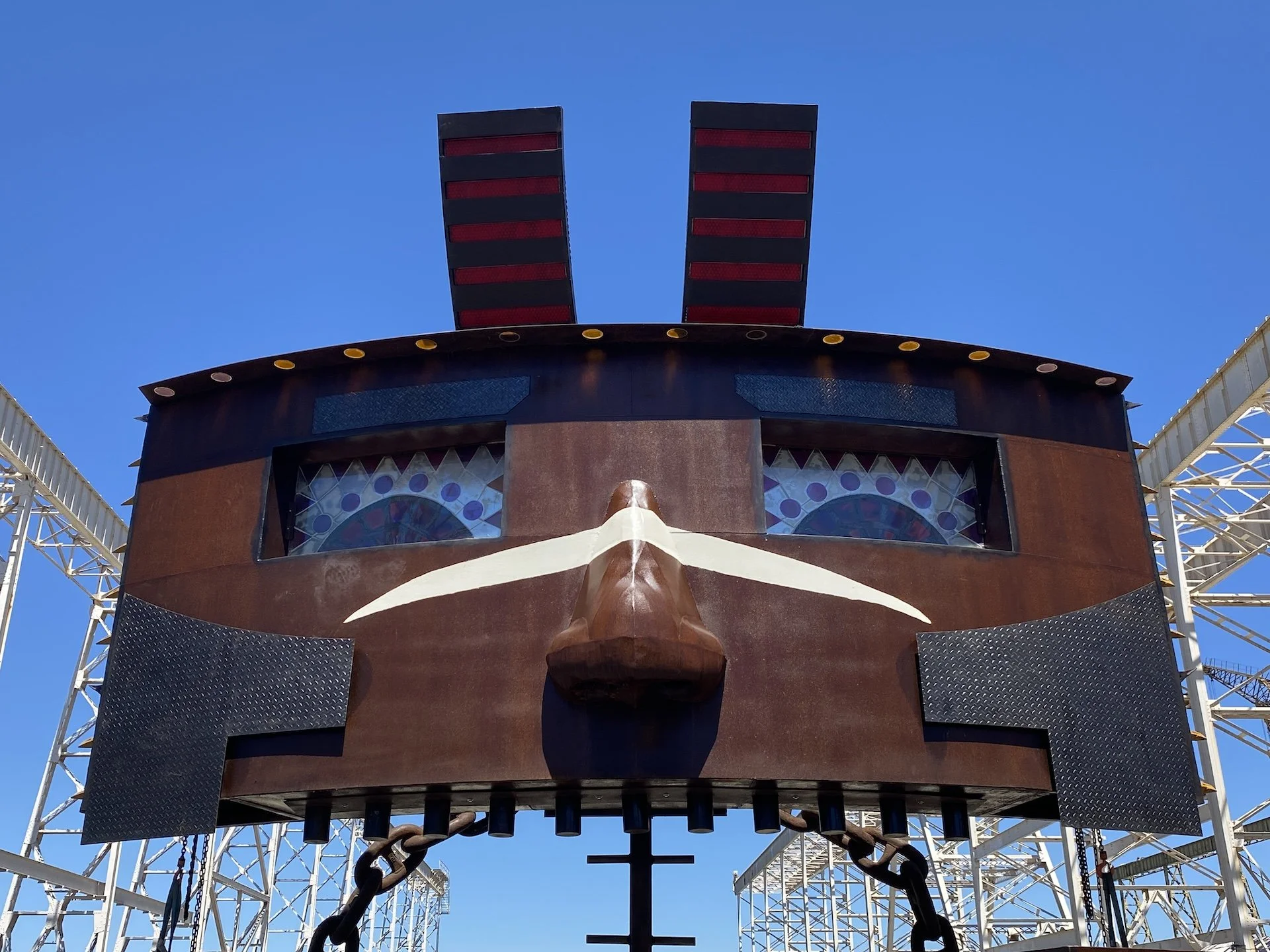 Close-up of a carnival ride face with a large animated face, including eyes, nose, and mouth, against a clear blue sky, with part of a roller coaster visible on the sides.