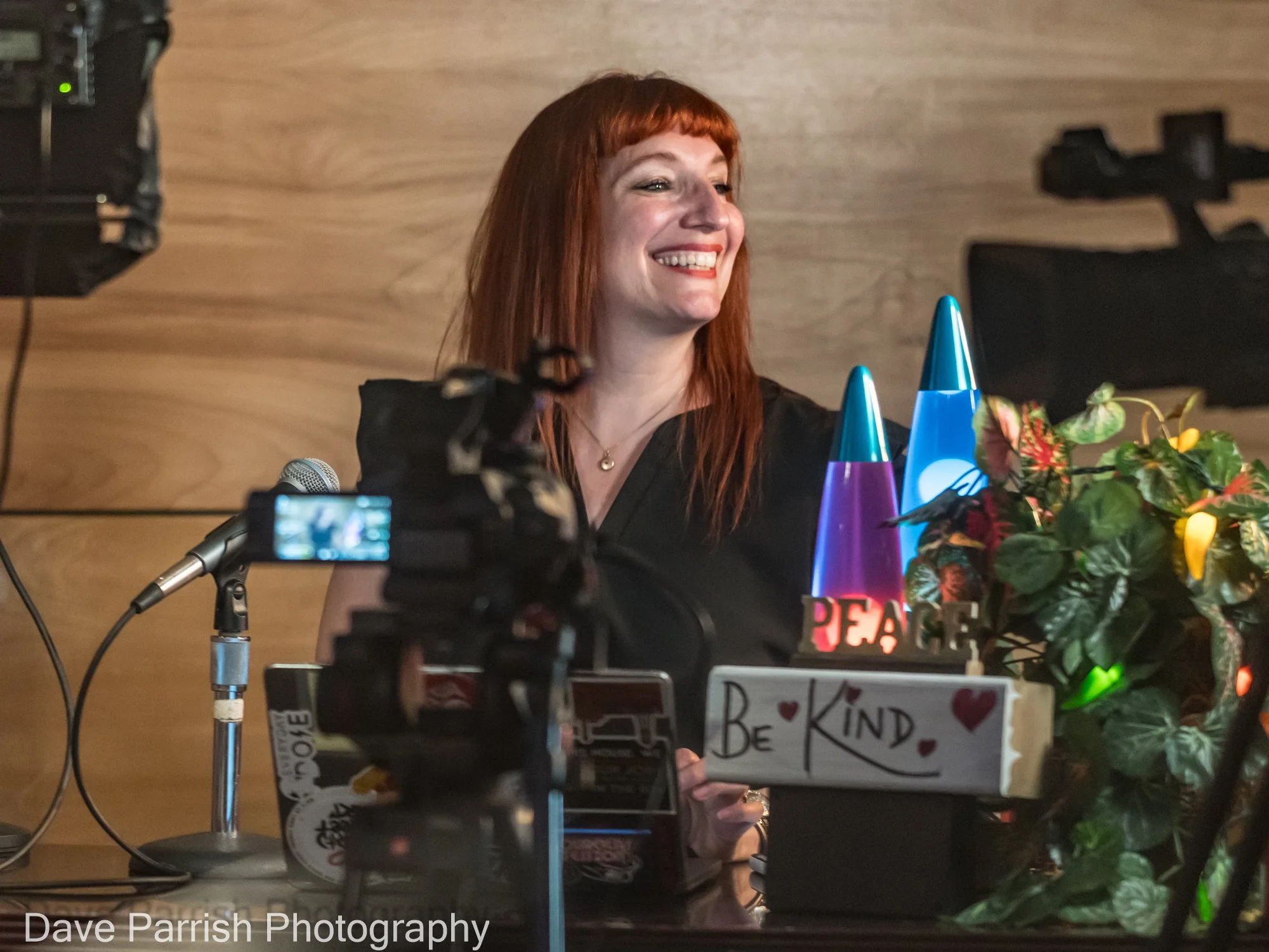 A woman smiling behind a desk, with a camera in front of her.