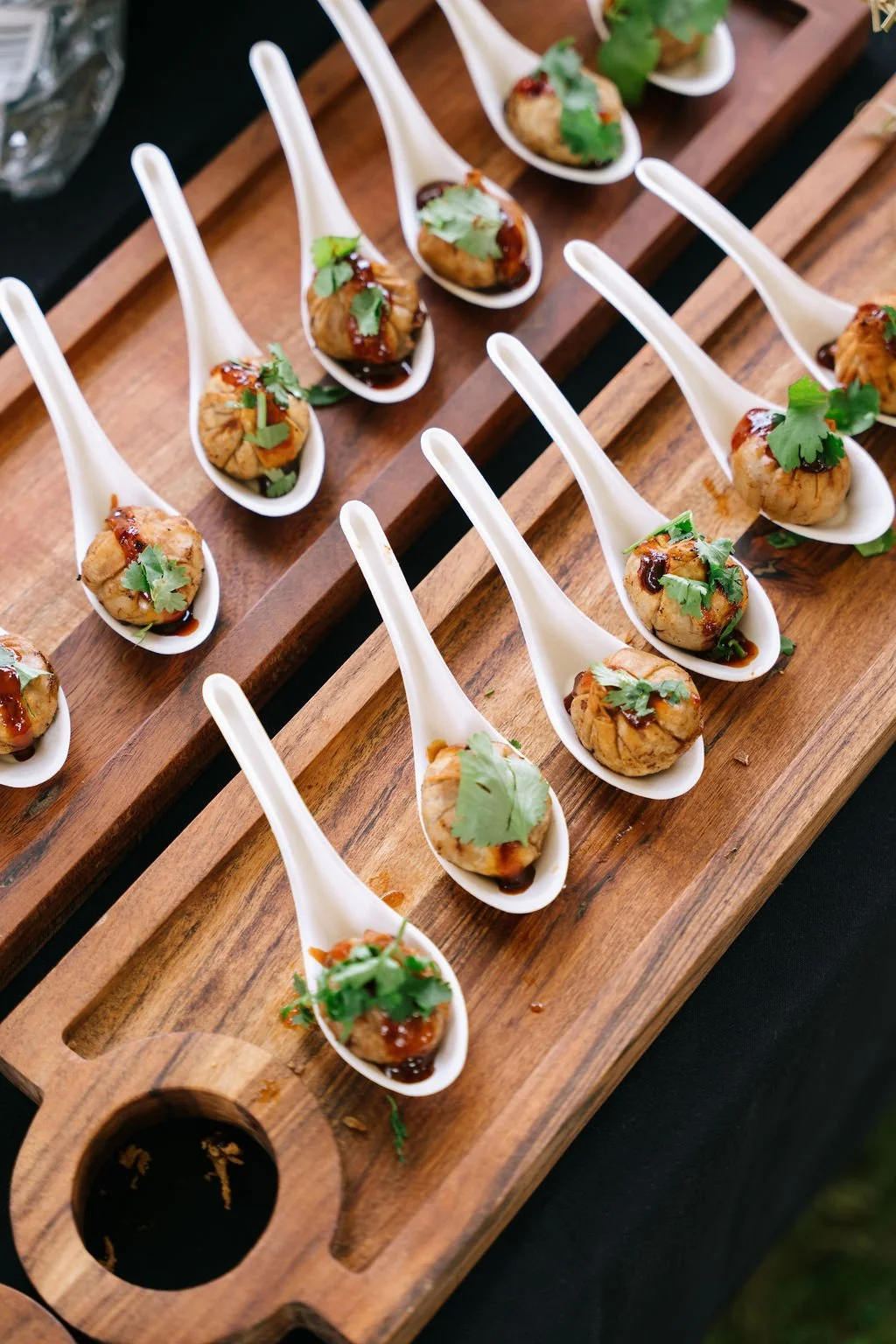 Multiple bite-sized appetizers served on white ceramic spoons arranged on a wooden tray, garnished with cilantro and drizzled with sauce.