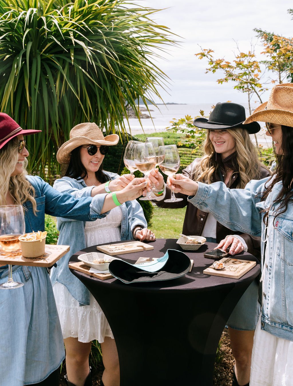 A group of women outdoors, wearing sun hats and sunglasses, raising wine glasses in a toast near a table with food, greenery, and water in the background.