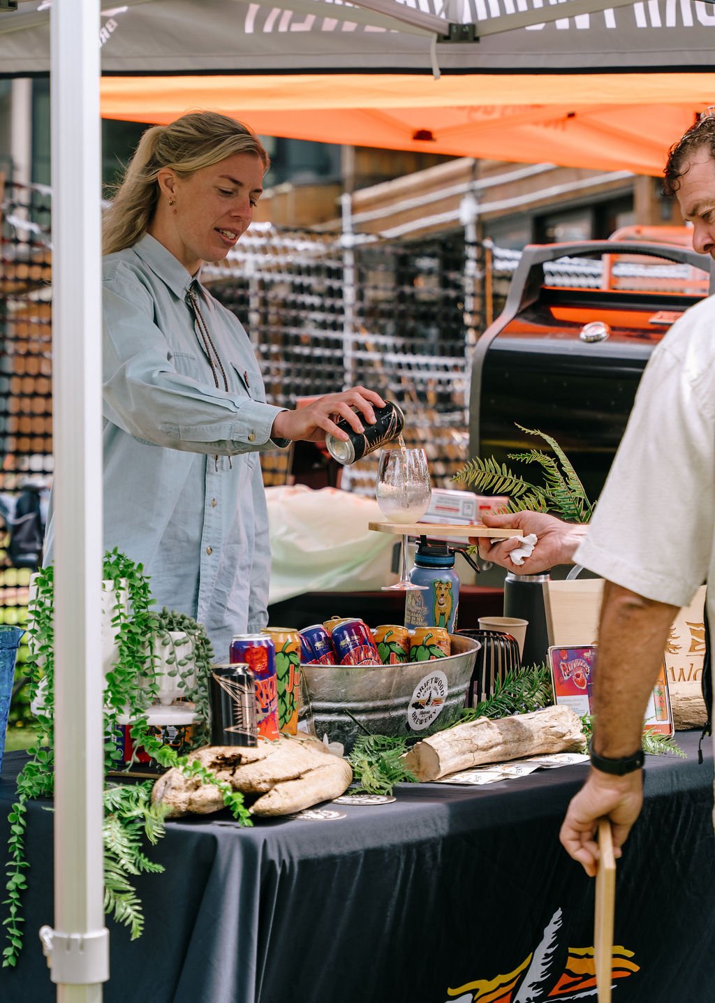 A woman is at a market stall pouring a drink into a glass for a man holding a tray. The stall has cans of beverages, plants, and rocks, with a black tablecloth. The background features a wood wall and orange tent.