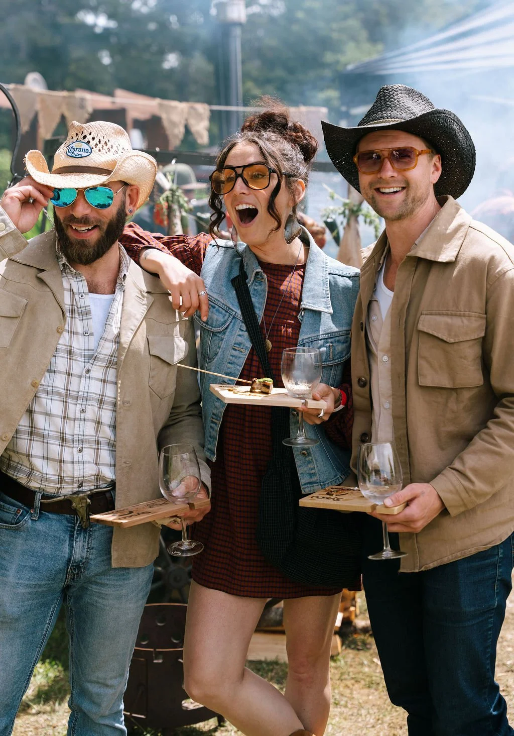 Three friends in sunglasses and cowboy hats smiling at a backyard gathering, holding wine glasses and a platter of food.