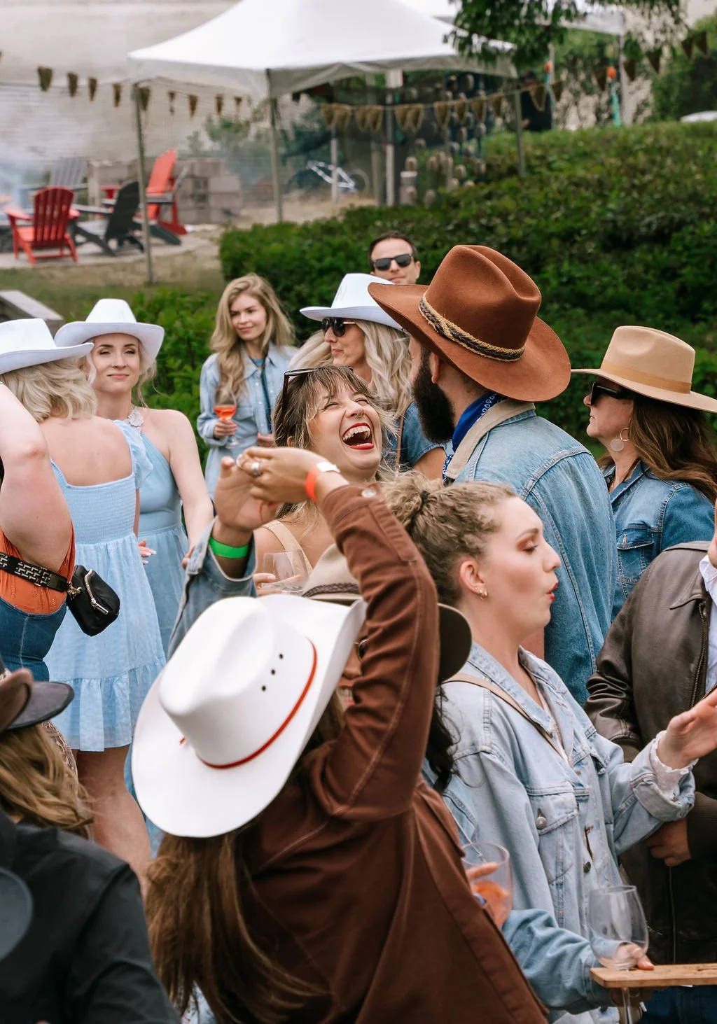 Group of people at an outdoor gathering, many wearing cowboy hats, laughing, talking, and enjoying drinks in a garden with a tent and outdoor furniture in the background.