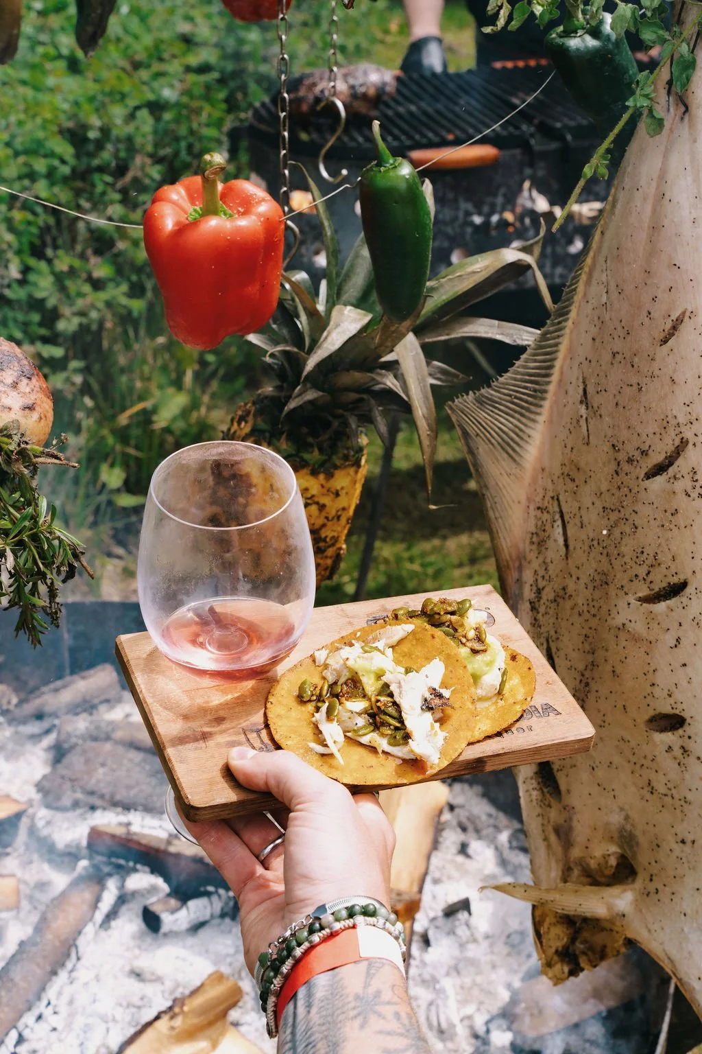 Hand holding a wooden board with two tostadas topped with cheese and green peppers, and a glass of pink wine. In the background, a red bell pepper, a green jalapeño, and a pineapple are hanging from a plant, with a grill in a green outdoor setting.