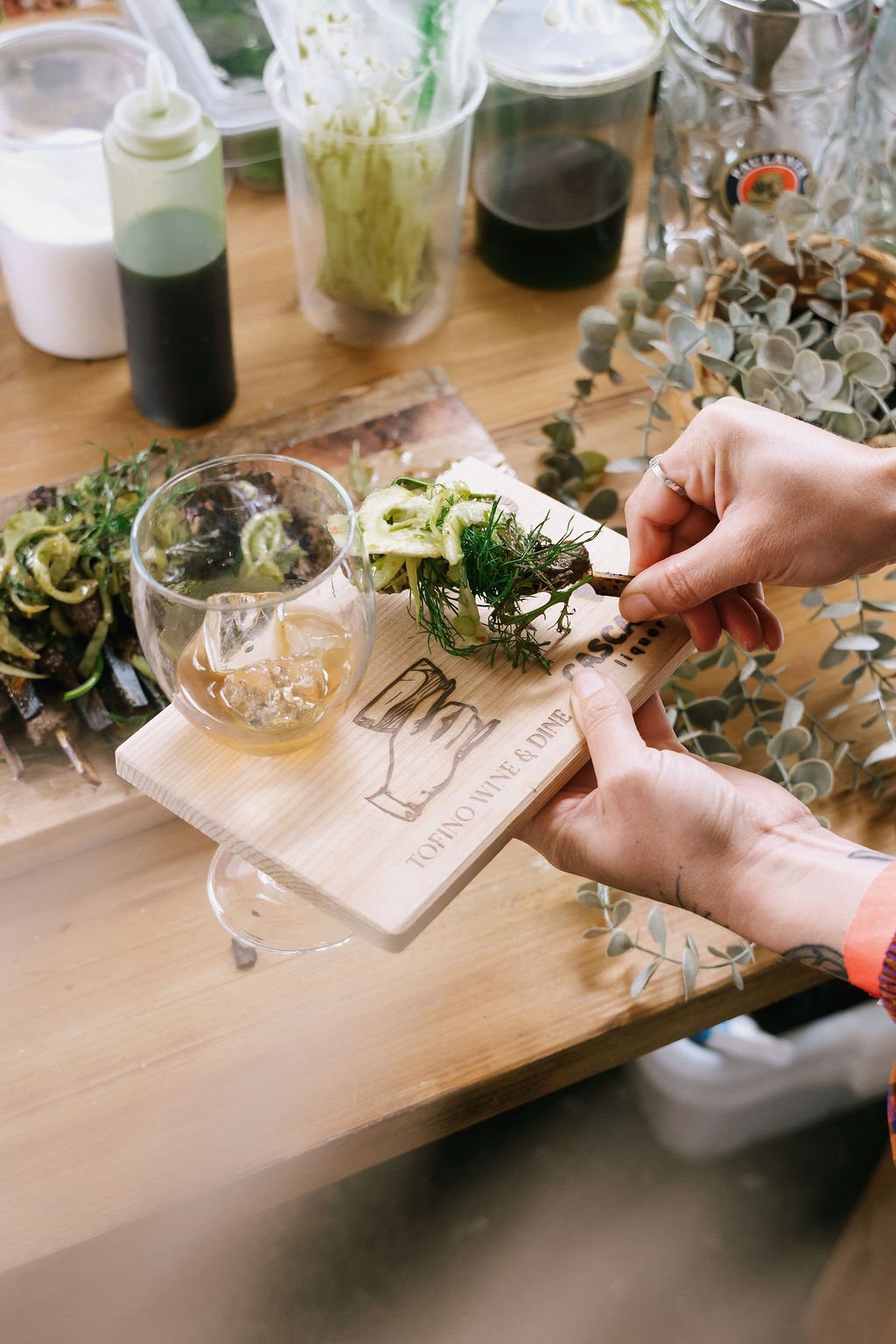 Person holding a wooden serving board with a small salad, a glass of wine with ice, on a wooden table with various drinks and plants in the background.