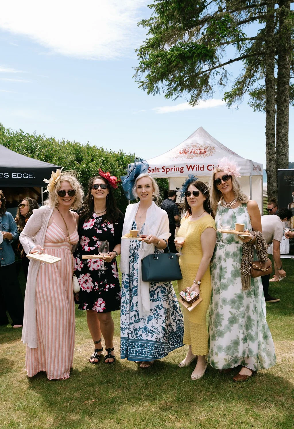Five women dressed in spring attire with fascinators, standing outdoors at a festival, holding wine glasses and plates, with tents and trees in the background.