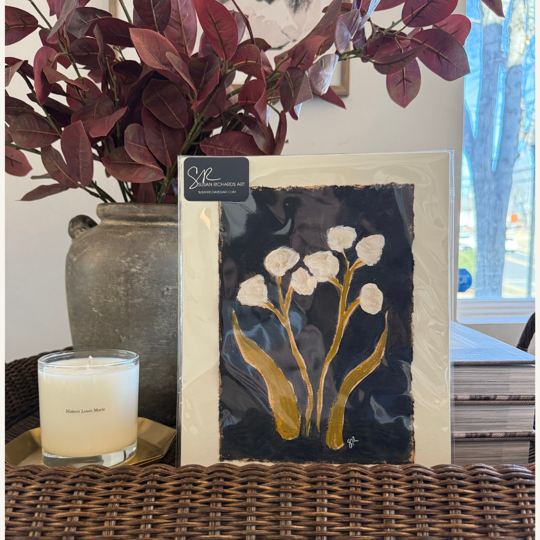 A framed painting of white flowers with yellow stems and leaves, positioned on a wicker surface. To the left, a lit candle in a glass holder labeled 'Maison Louis Marie'. Behind the painting, there is a large vase with purple and green leaves, and stacks of books on the right side. A window with blue icicle decorations can be seen in the background.
