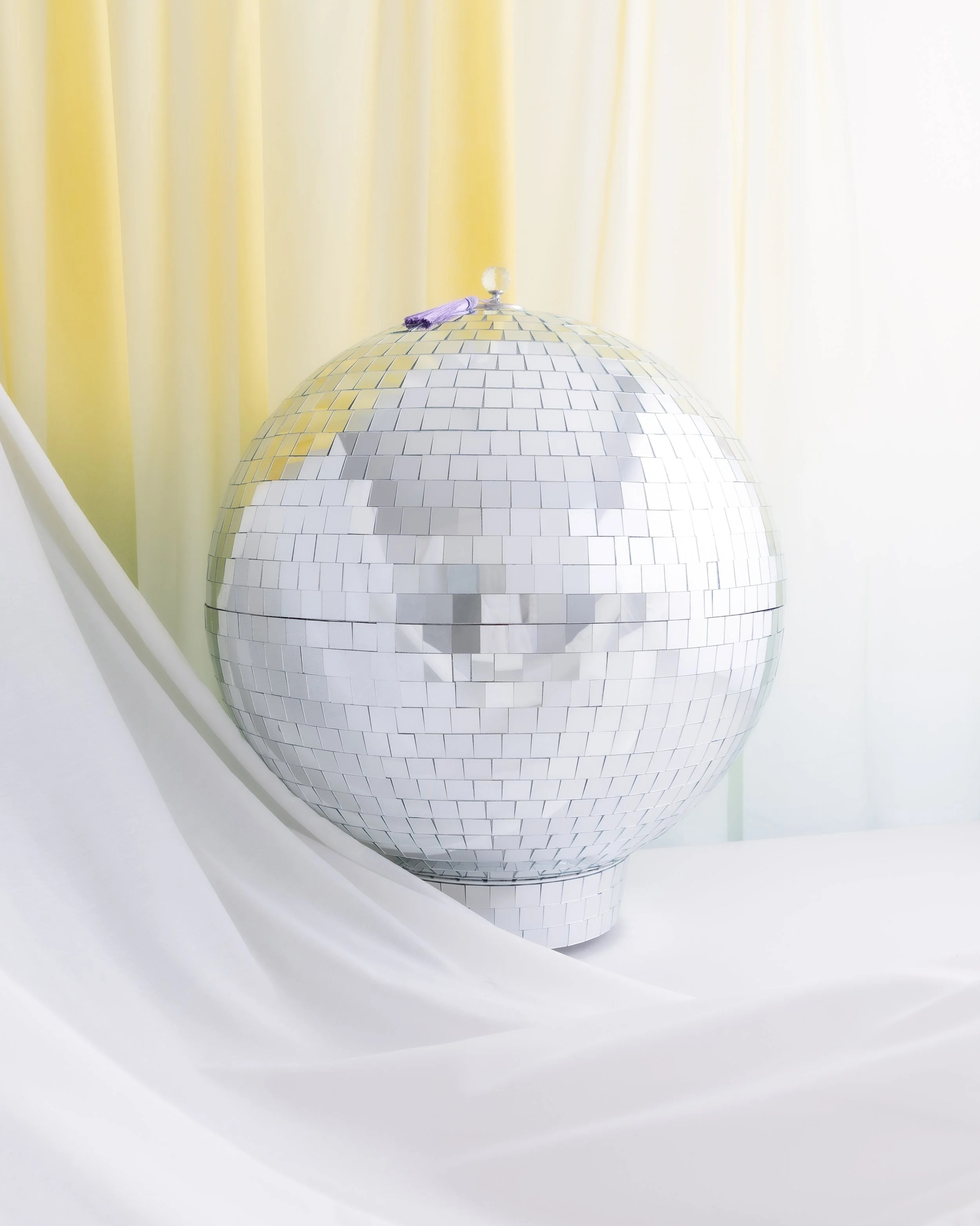 A silver disco ball with shiny tiles on a draped surface and a yellow curtain backdrop.