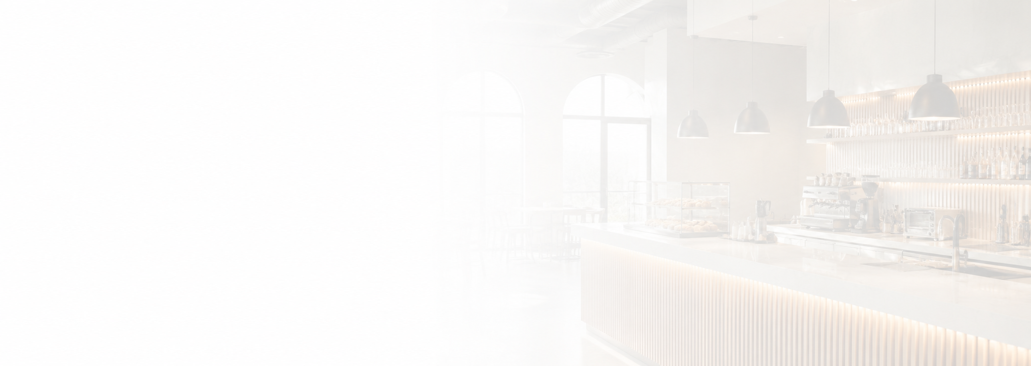Bright cafe interior with white walls, large arched windows, pendant lights, and a counter with a glass display, coffee machines, and shelves with bottles and jars.